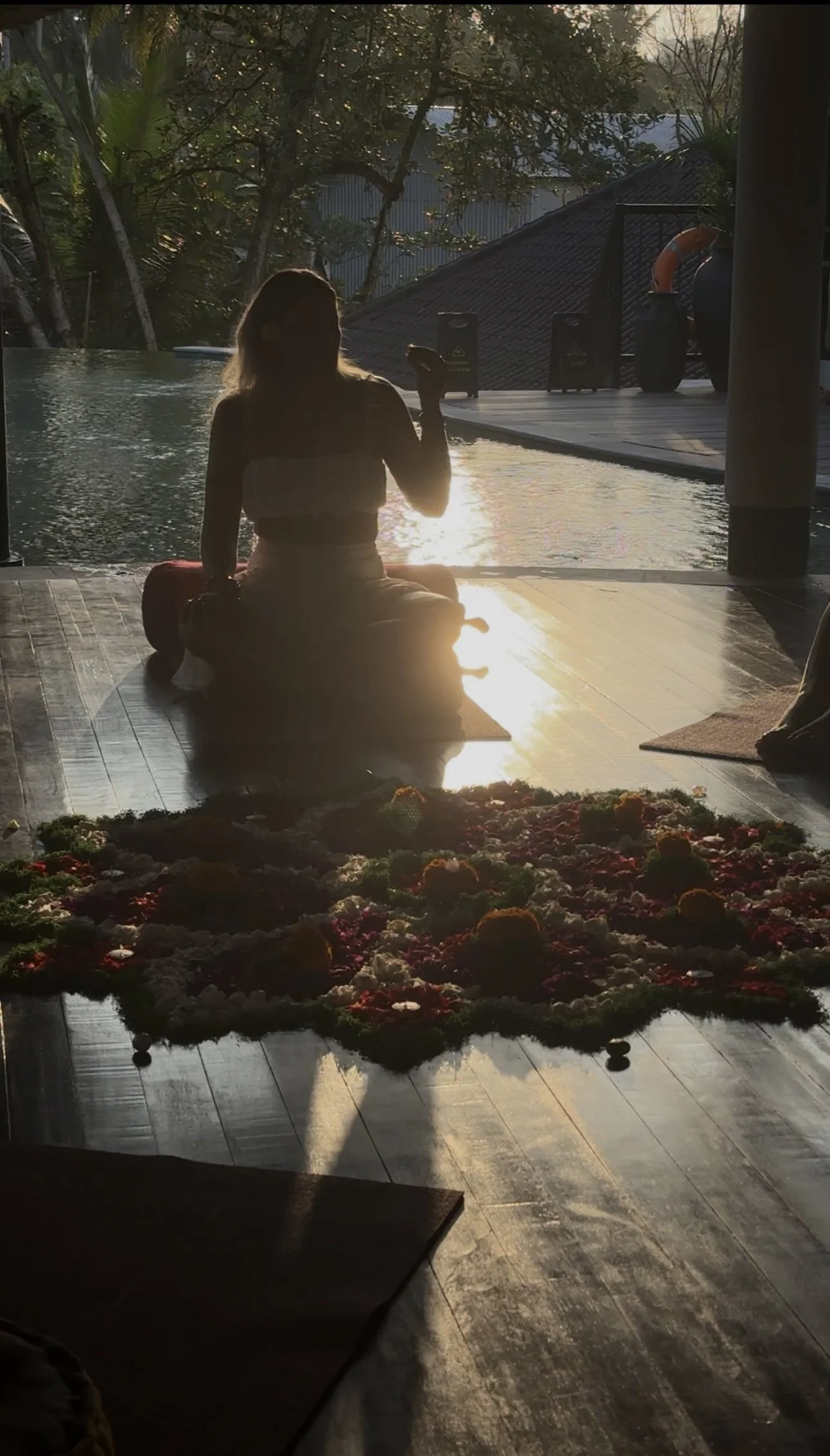 Backlit woman sitting in meditation beside a resort pool at sunset, with colourful flowers in the foreground and tropical trees behind her
