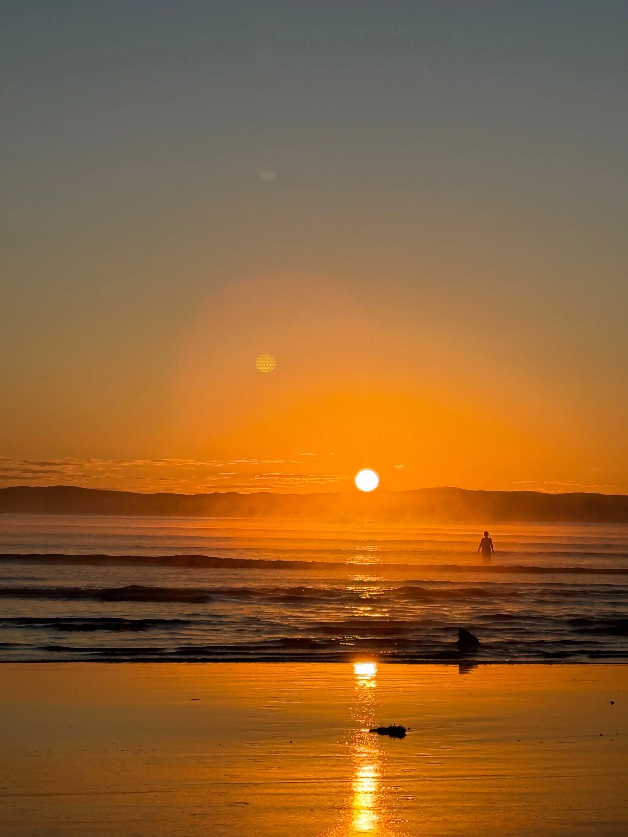 Vibrant orange sunset over ocean waves, with a silhouetted person wading in the water and sunlight reflecting on the wet beach.