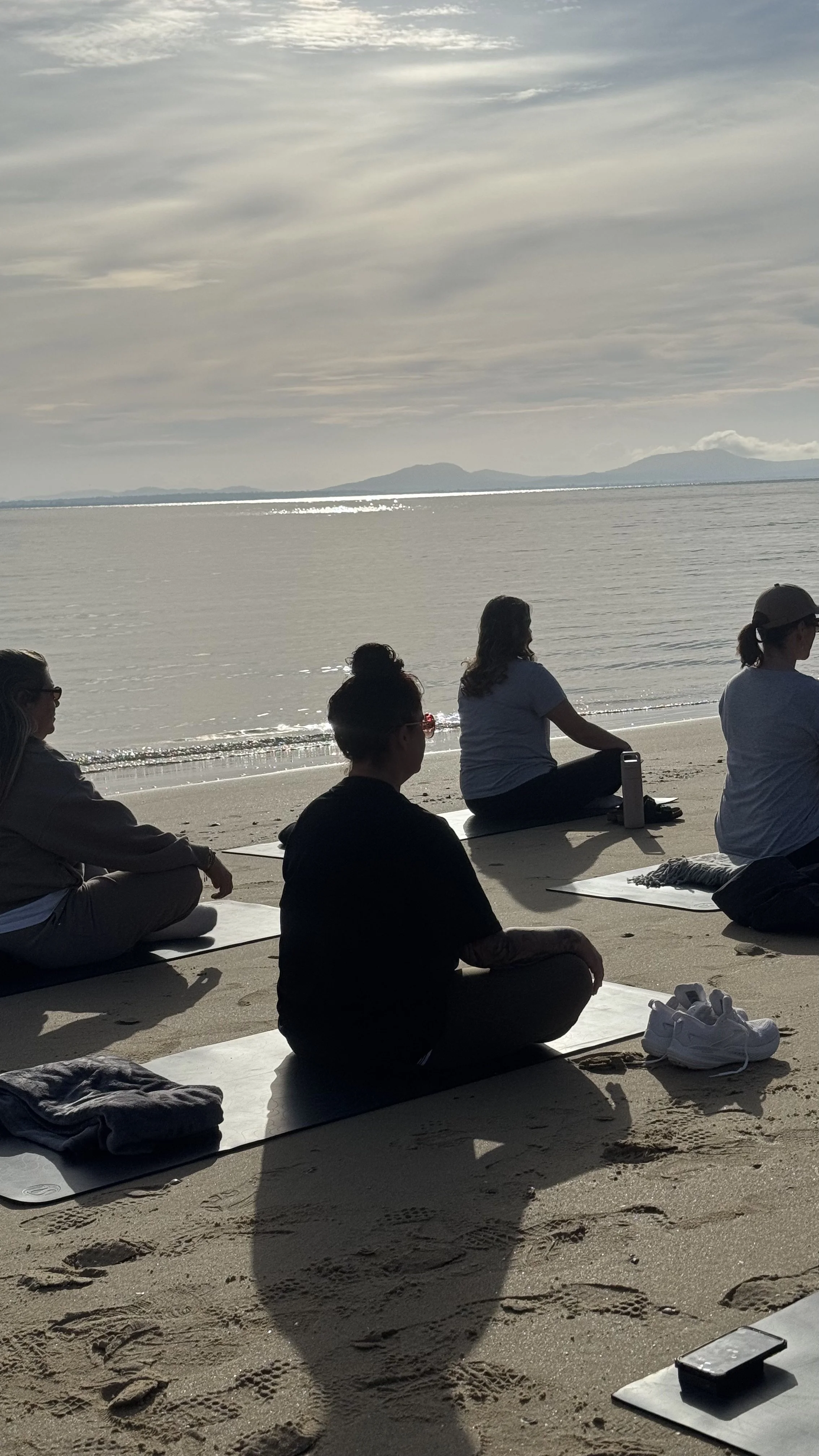 People sitting on yoga mats at a beach meditation class, with calm water and mountains in the background.