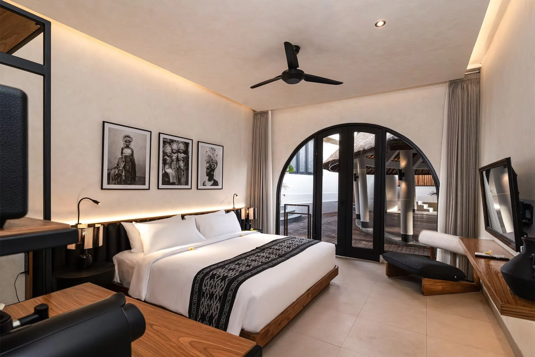 Luxury tropical hotel bedroom with a king bed, black ceiling fan, arched glass doors, framed artwork, and private courtyard