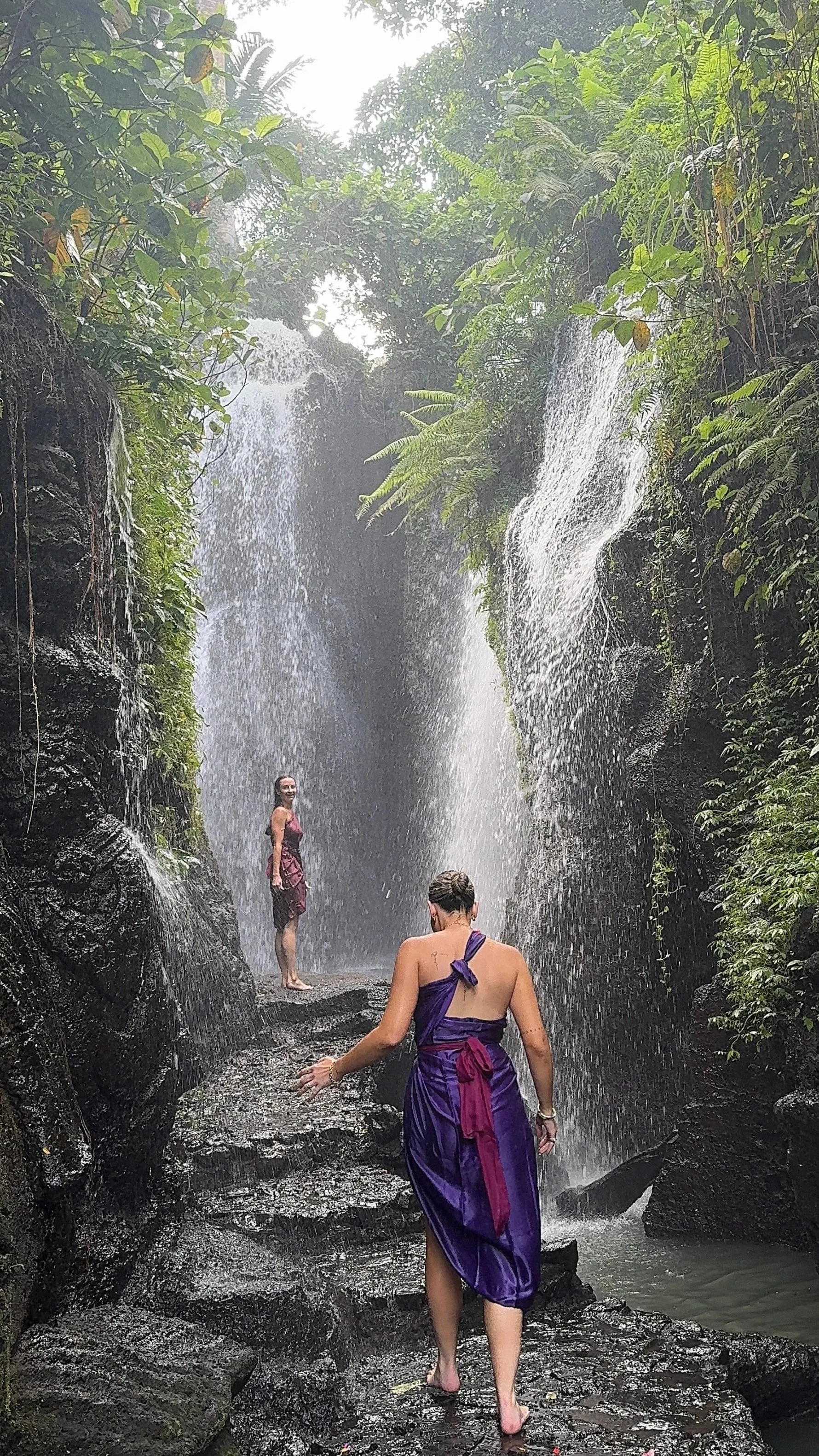 Two people explore a lush tropical waterfall, with one woman standing beneath the cascading water and another in a purple dress in the foreground.