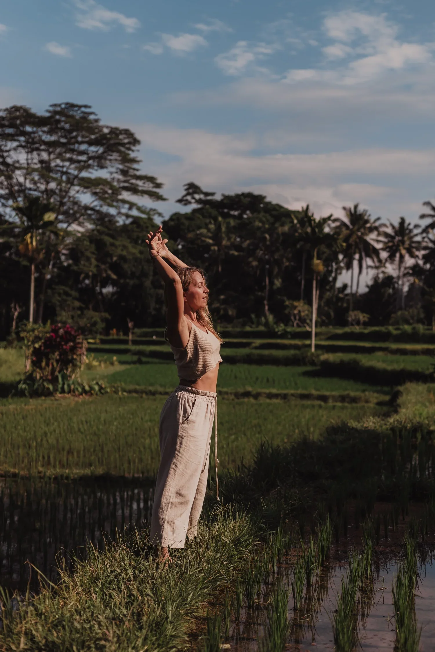 Woman stretching with raised arms beside a reflective rice paddy in a tropical landscape