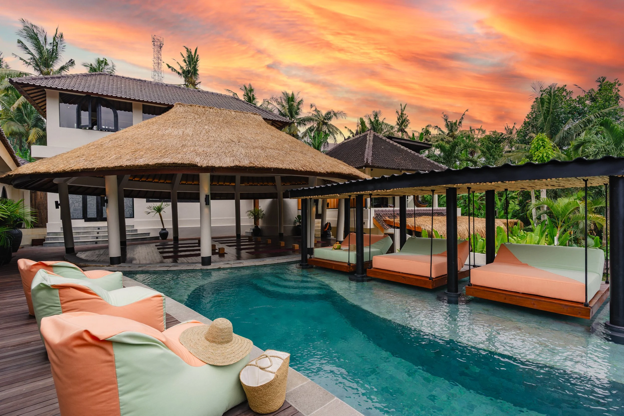 Tropical resort villa with a thatched-roof pavilion, turquoise pool, floating lounge beds, palm trees, and a vivid sunset sky