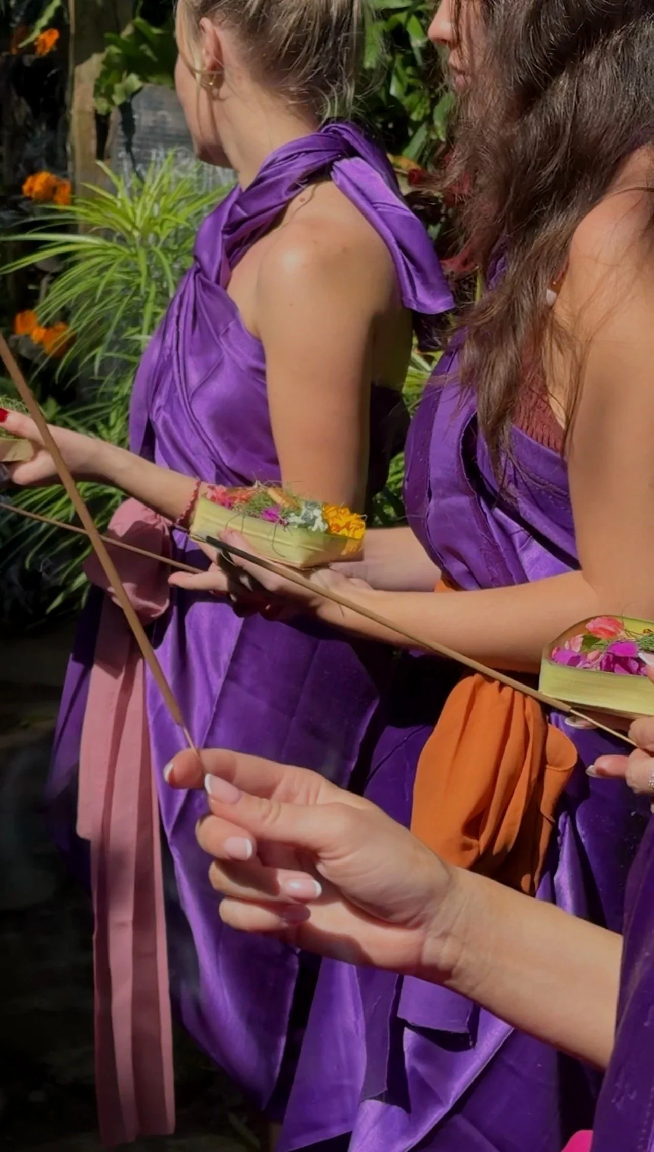 Women in purple dresses hold flower-filled palm-leaf offerings and incense sticks in a tropical garden.