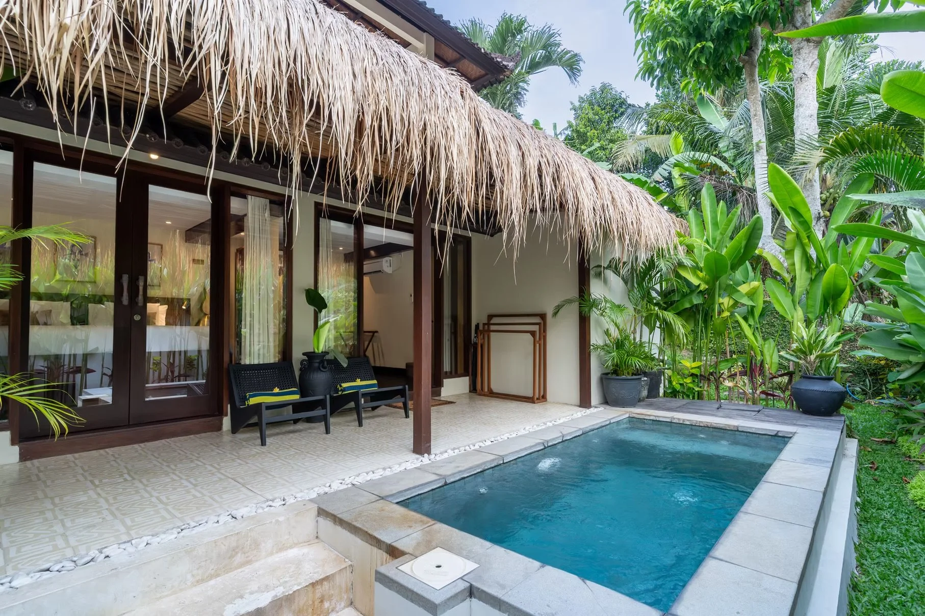 Tropical villa with a thatched-roof patio, lounge chairs, lush greenery, and a private swimming pool.