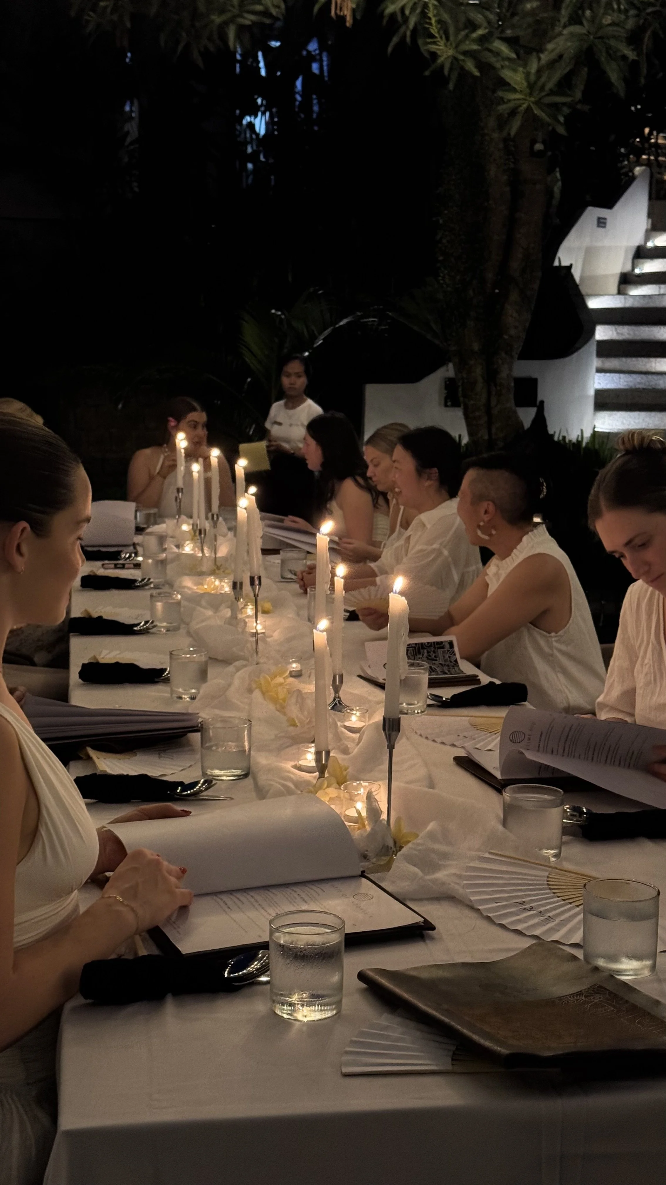 Guests dine at a candlelit outdoor restaurant table surrounded by tropical plants and a staircase.