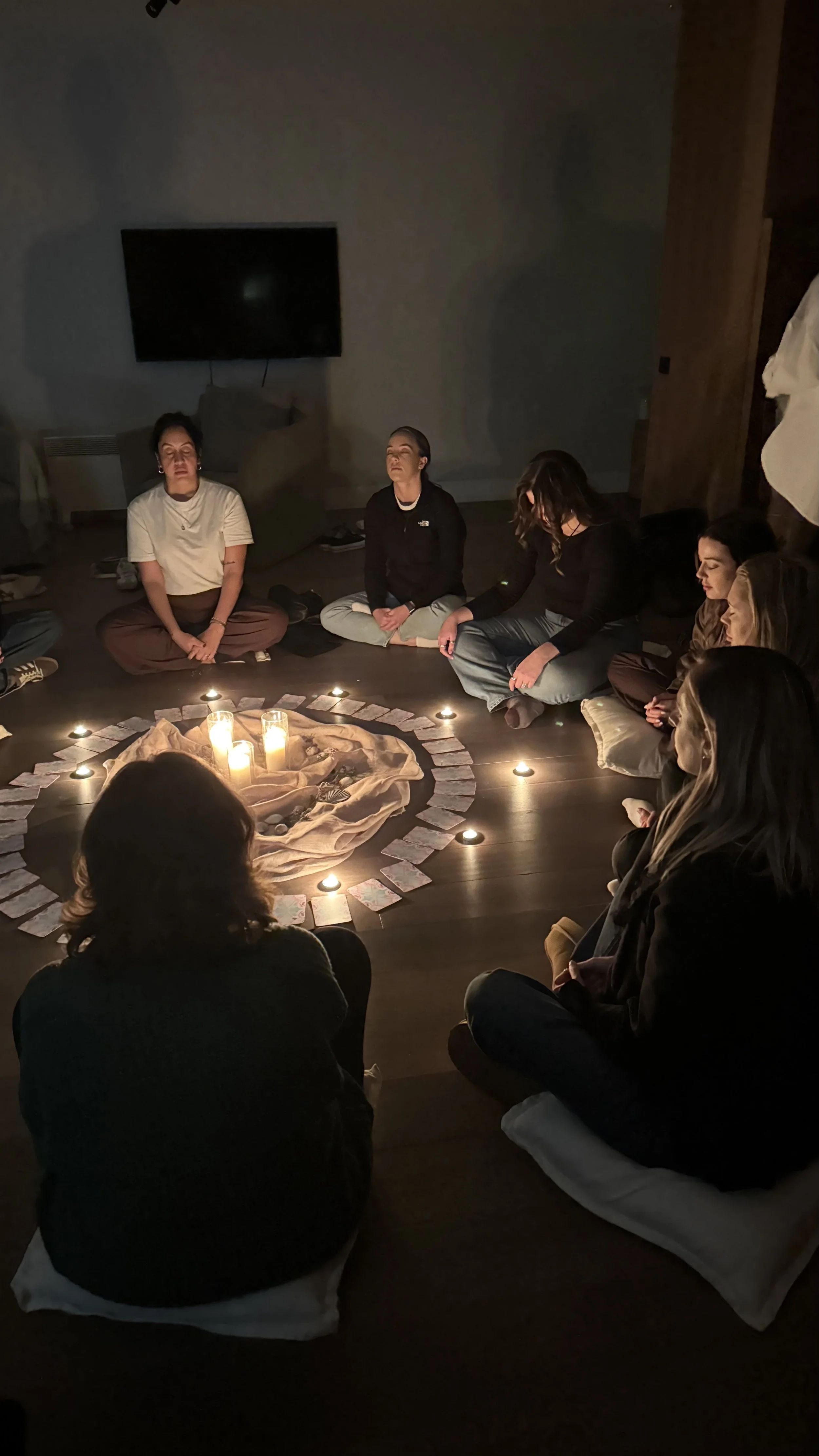 People sitting in a candlelit circle around cards and a central cloth-covered arrangement during a group meditation or ritual.