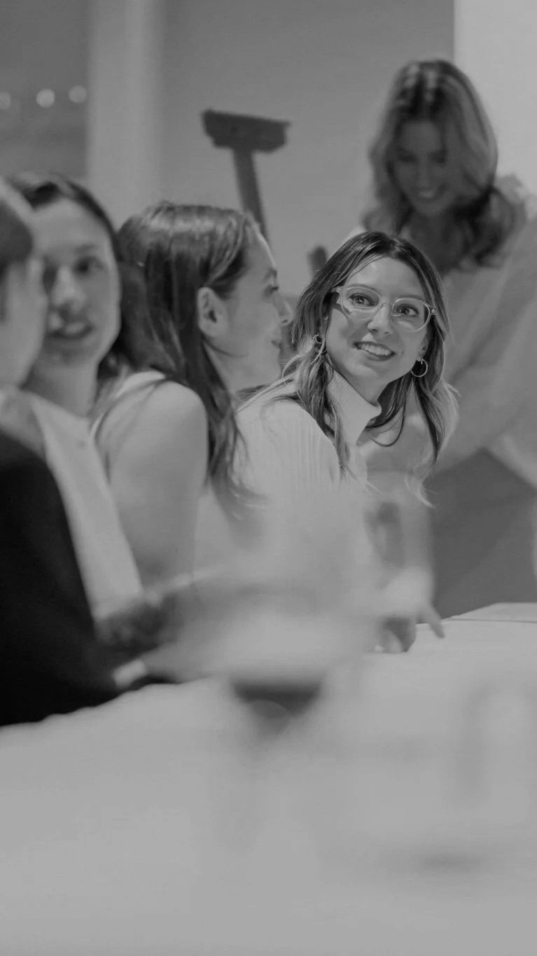 Black-and-white photo of women socializing around a table, with a smiling woman wearing glasses in the foreground.