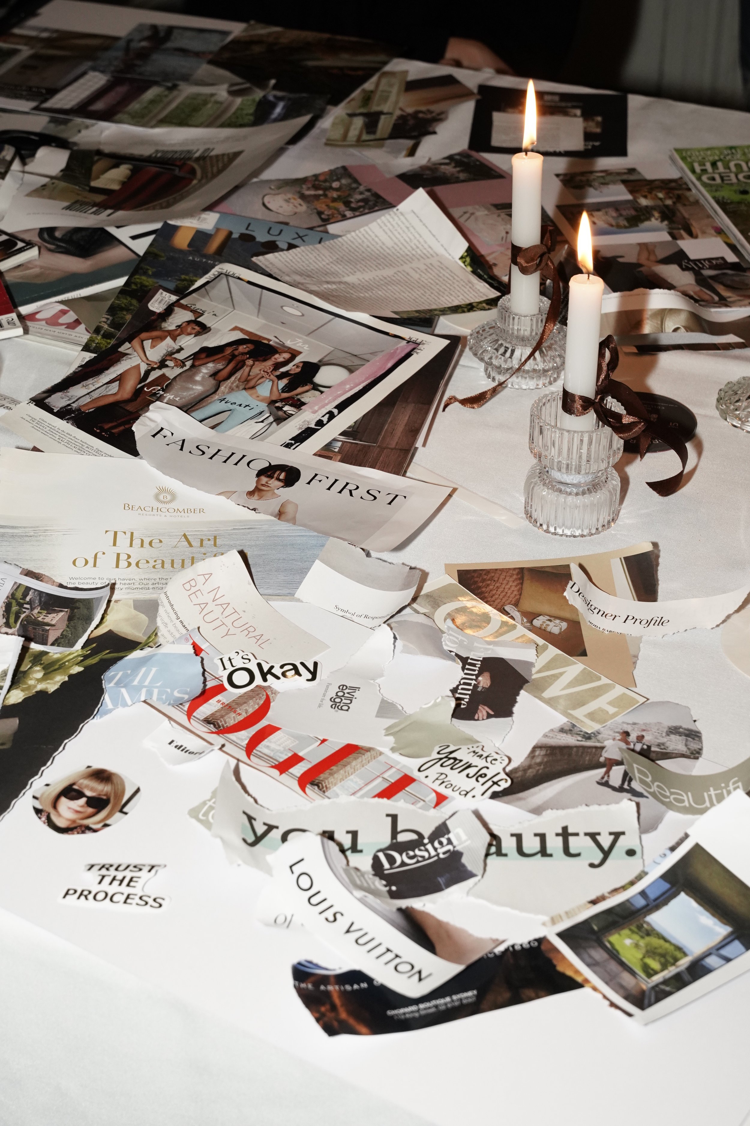Torn fashion and design magazine clippings spread across a table beside two lit white candles in crystal holders.