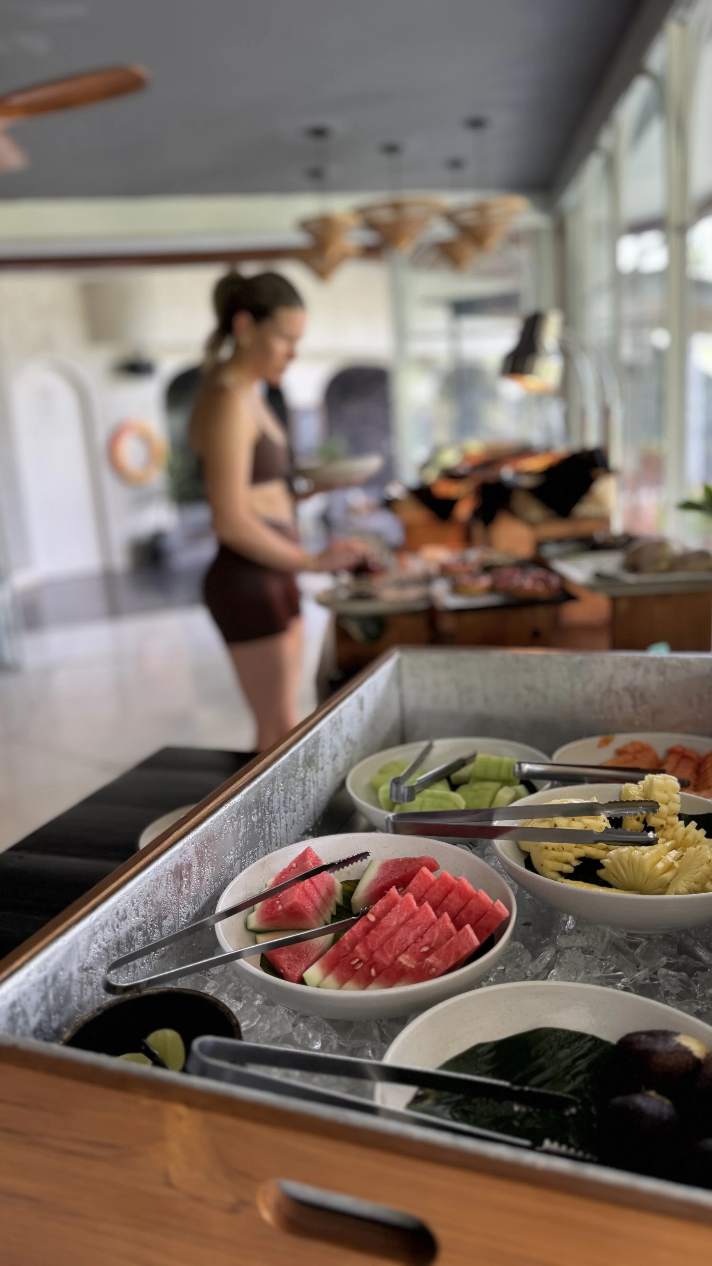 Blurry buffet display with bowls of sliced watermelon, pineapple, cucumber, and other fresh fruit, with diners in the background