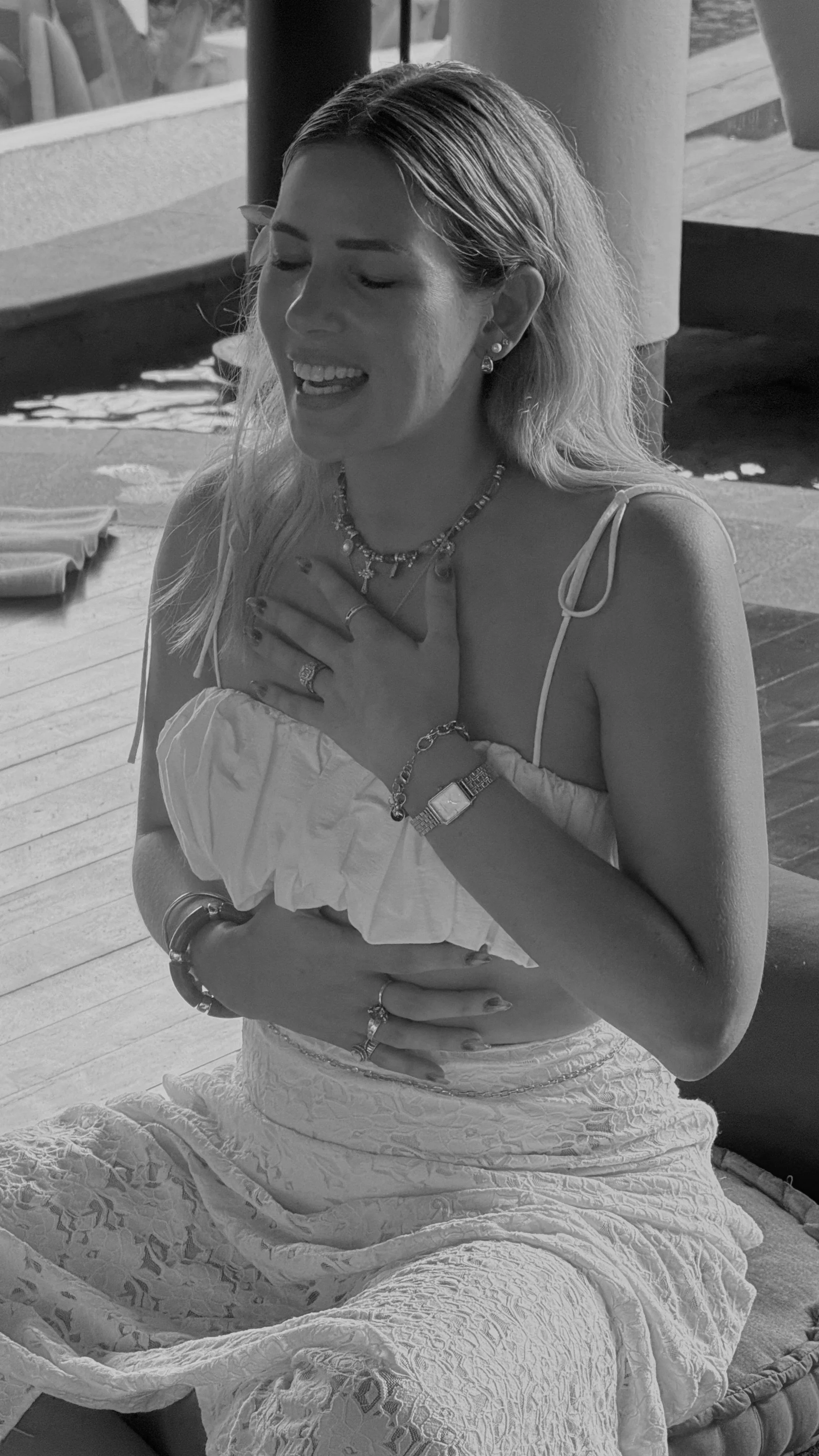 Smiling woman with long hair and jewelry sitting outdoors, wearing a sleeveless top and patterned skirt.
