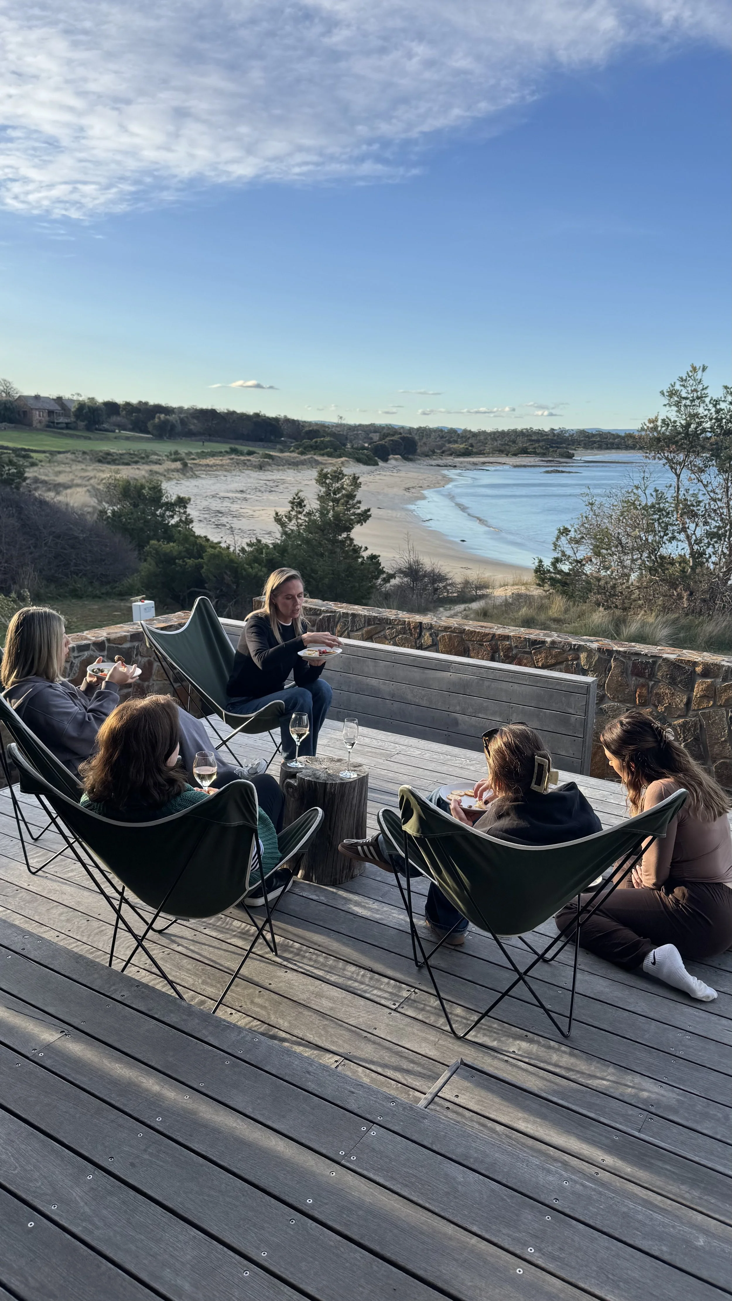 People gathered on a wooden deck enjoying food and wine overlooking a sandy beach and calm coastal inlet.