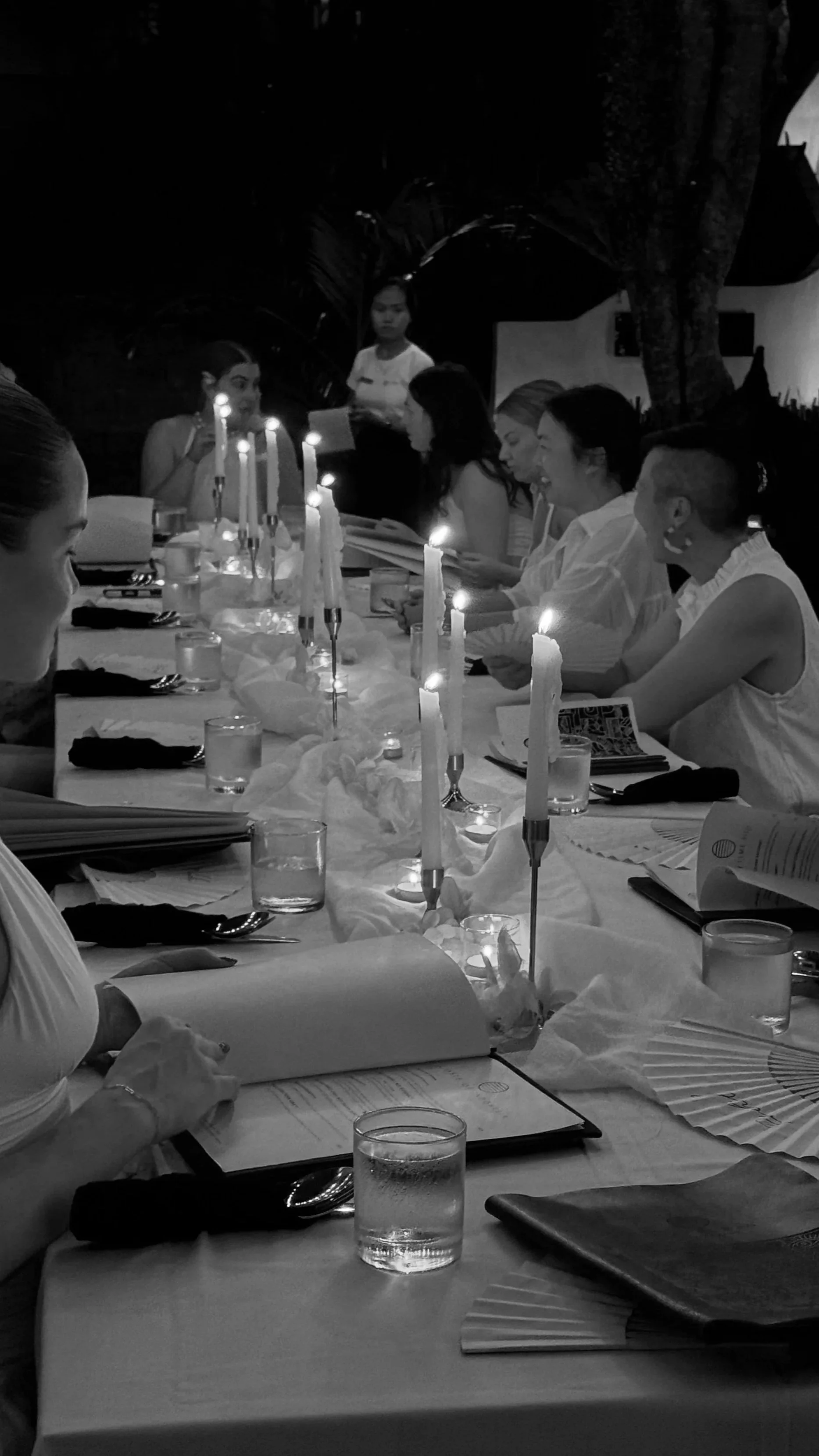 Guests dine at an outdoor Bali restaurant table illuminated by tall candles at night.