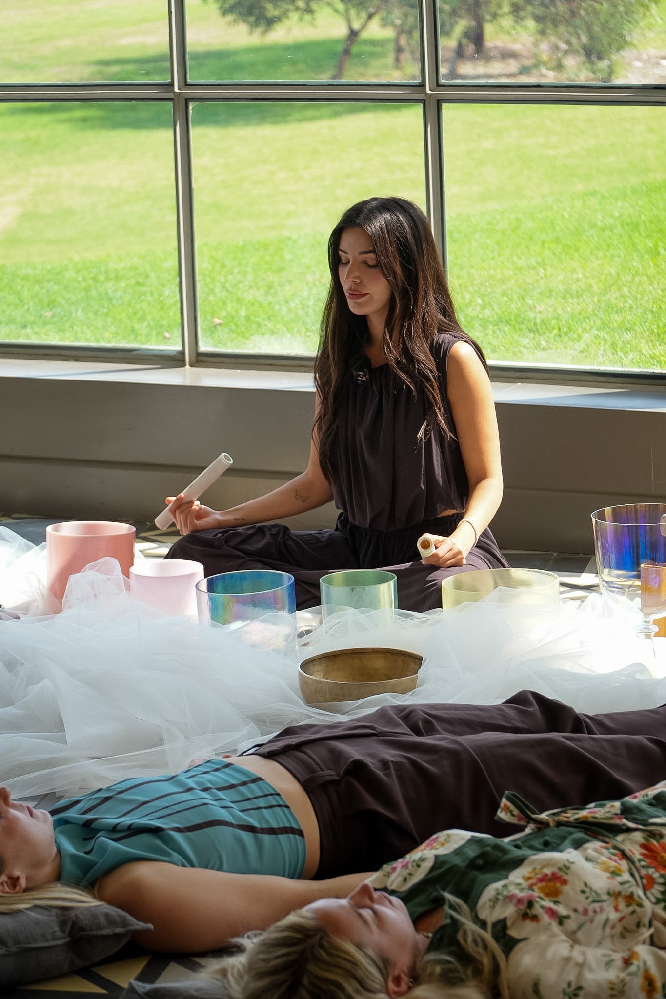 Woman holding a mallet beside colorful crystal singing bowls while another person relaxes on the floor in a sunny room
