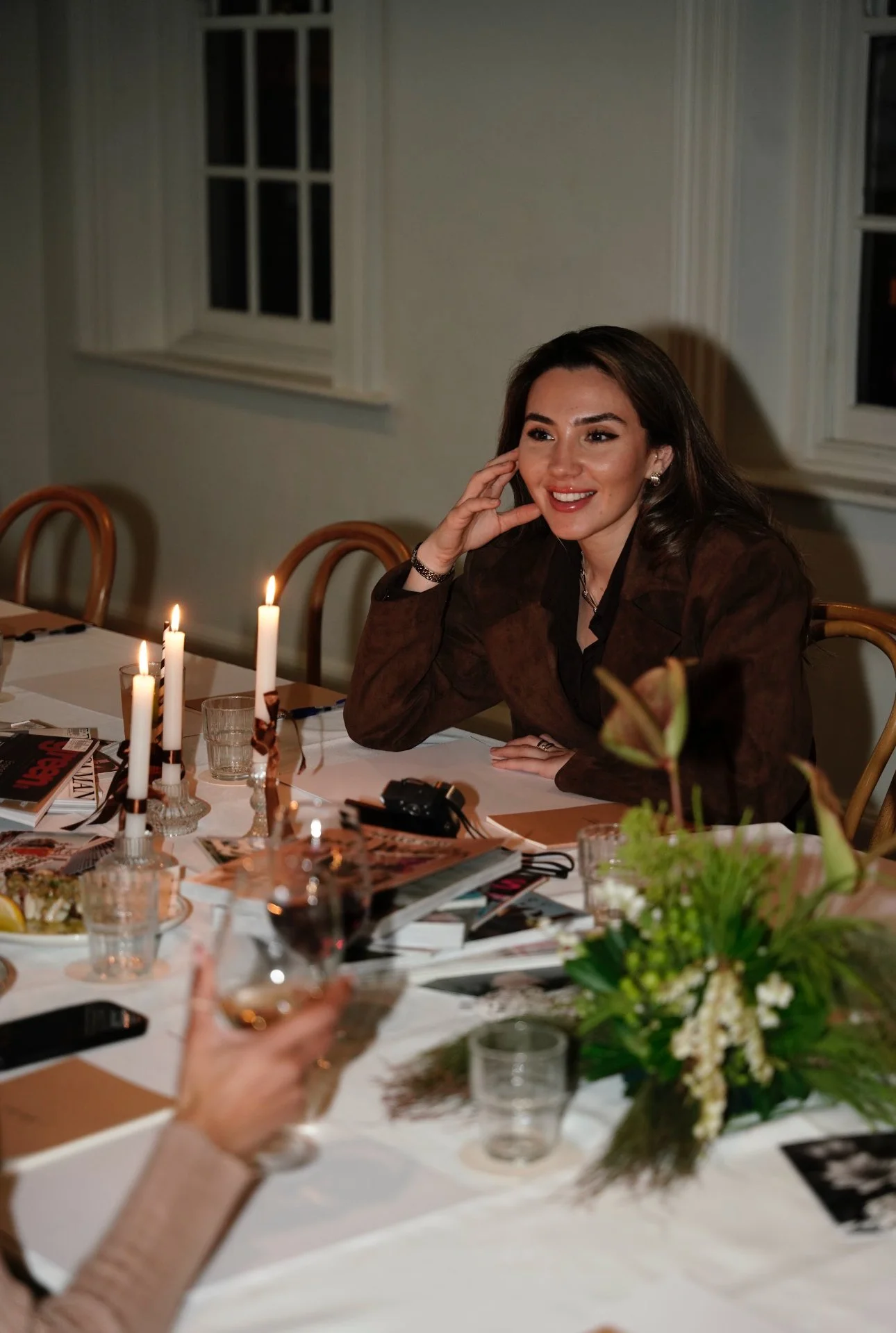 Smiling woman seated at a candlelit dinner table surrounded by magazines, drinks, and floral arrangements.