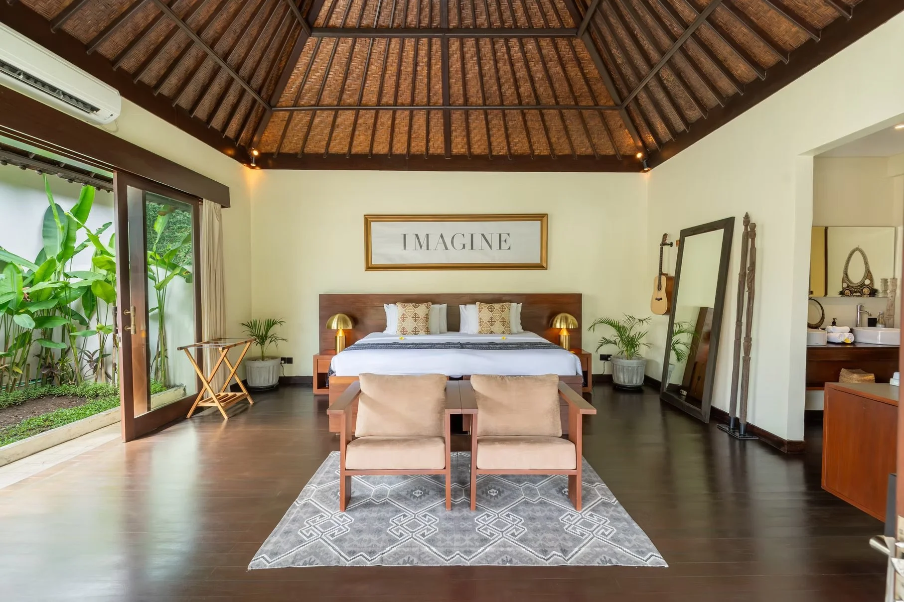 Luxury tropical bedroom with a thatched vaulted ceiling, king bed, patterned rug, lounge chairs, plants, and garden views