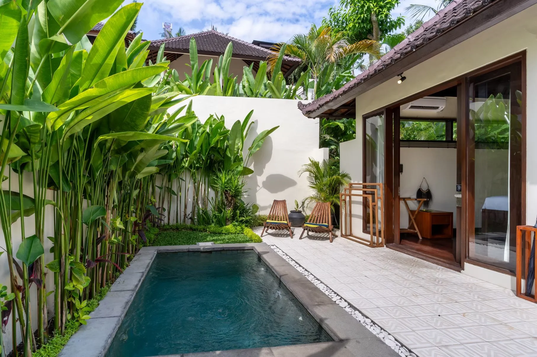 Tropical villa courtyard with a small swimming pool, lush banana plants, lounge chairs, and traditional tiled roofs.