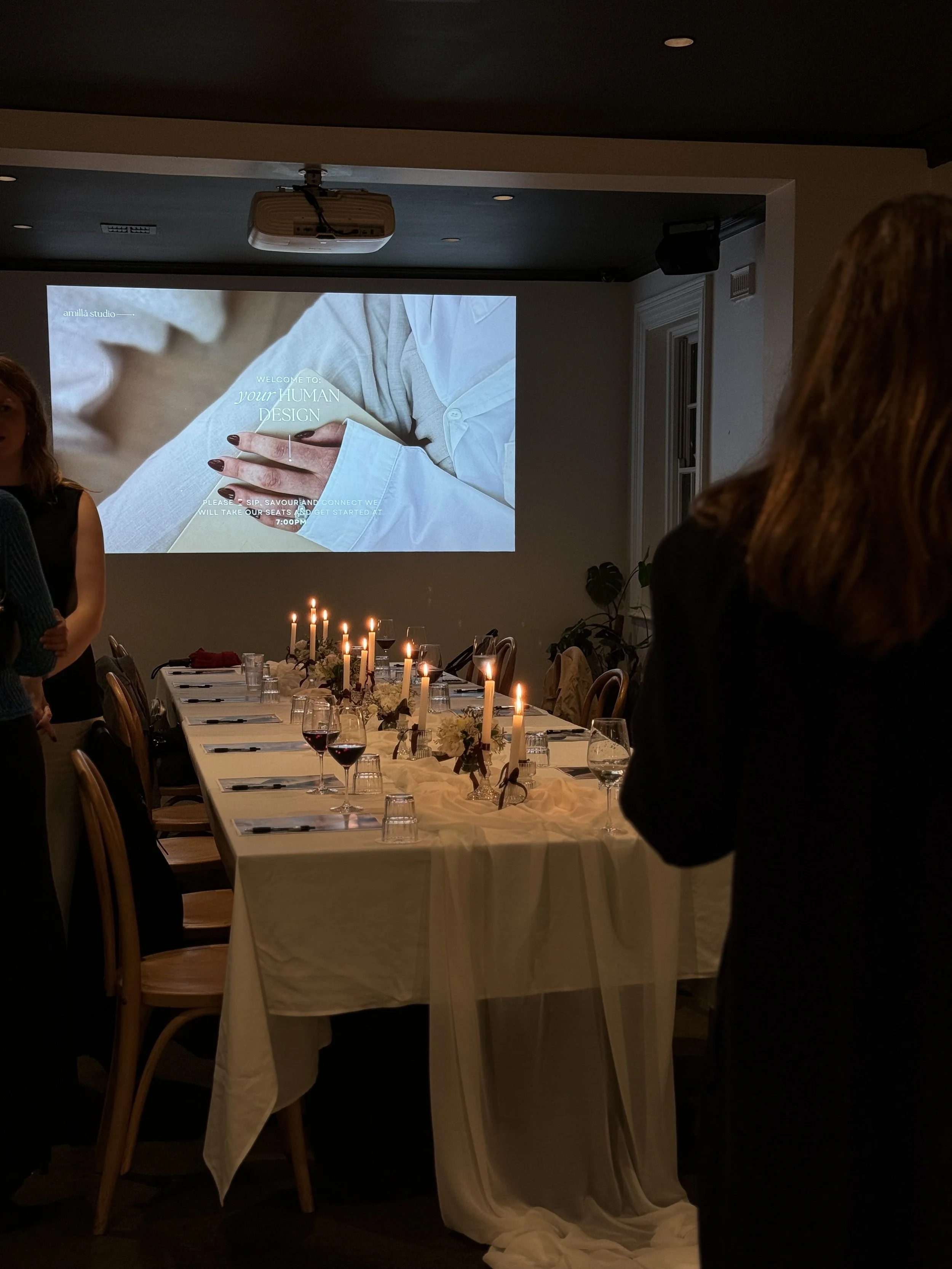 Candlelit dinner event with a long white-clothed table, wine glasses, floral centerpieces, guests, and a projected presentation.