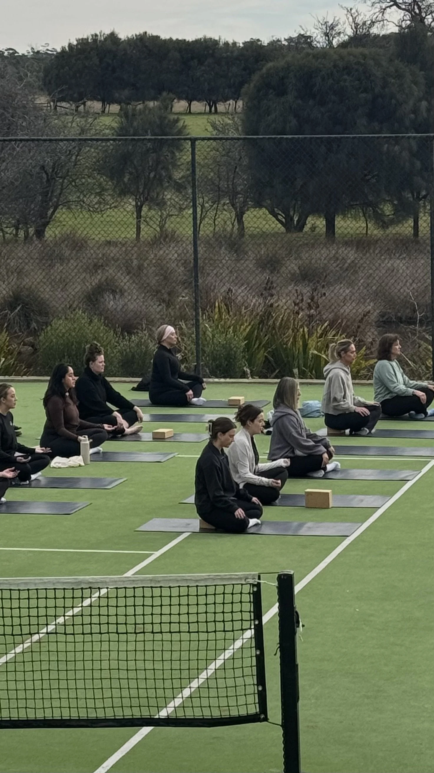 Group meditation class seated on yoga mats on an outdoor tennis court