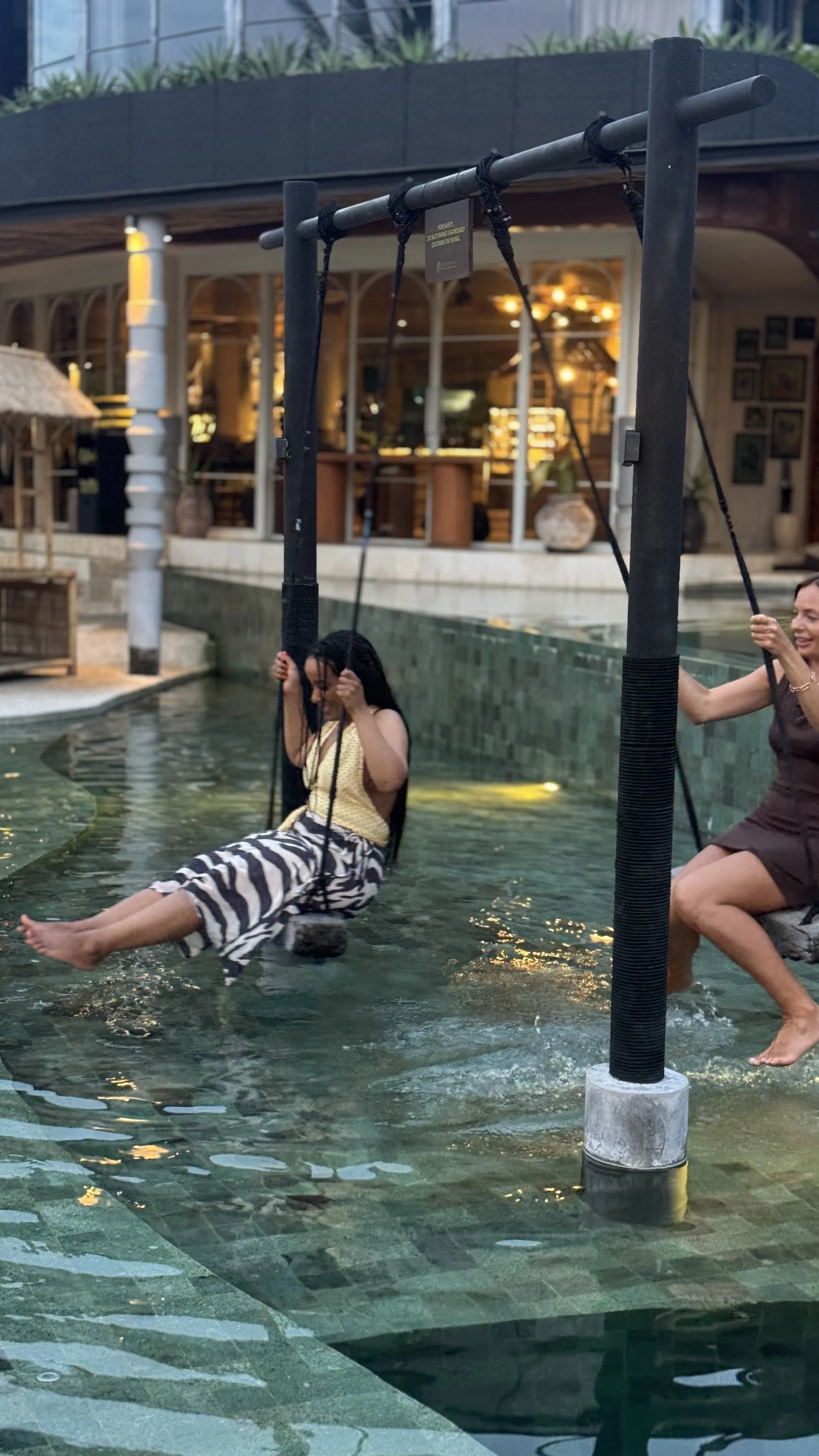 People swing from black ropes over a shallow indoor pool at an entertainment venue.