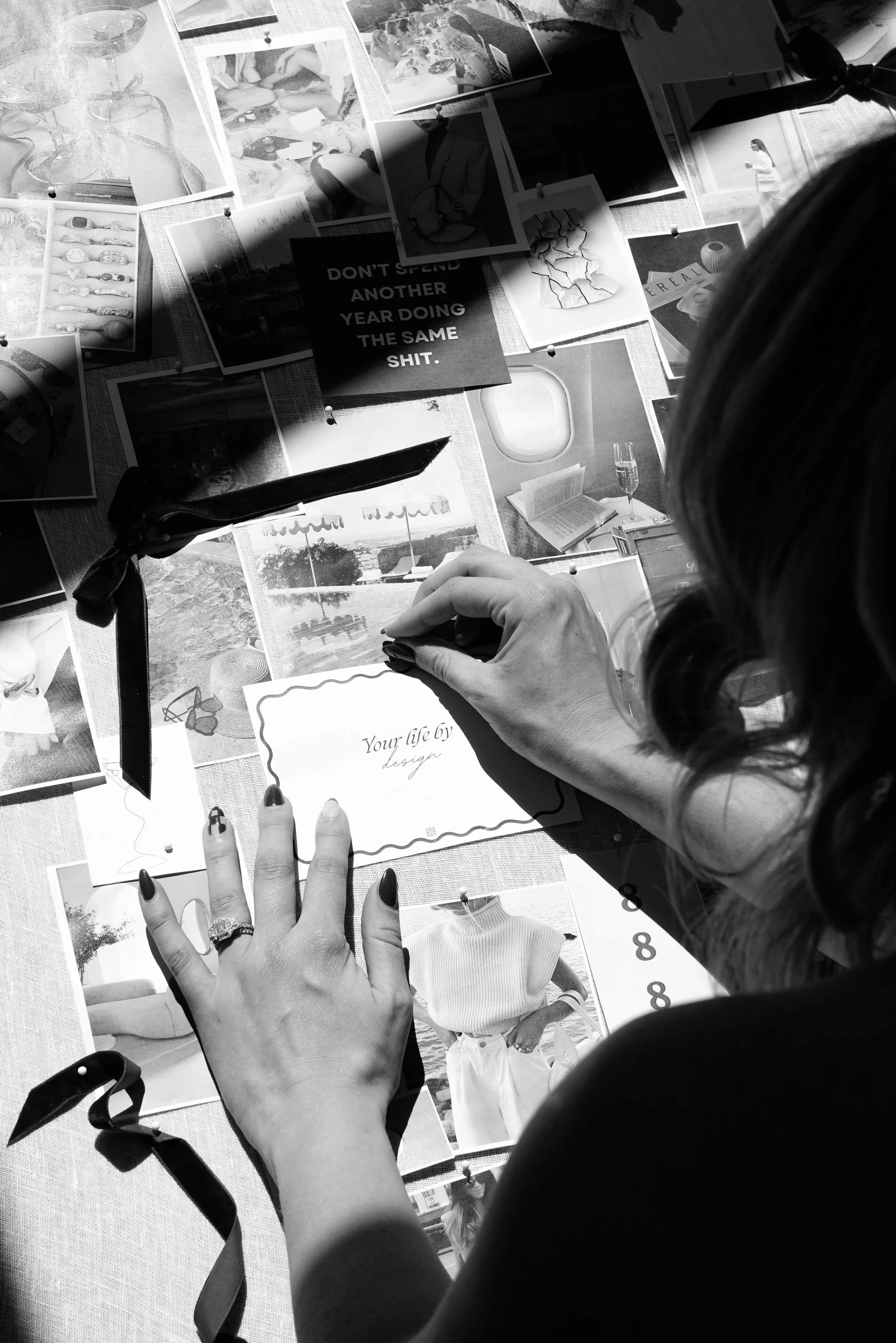 Black-and-white overhead view of a woman arranging pinned inspiration photos and typography on a mood board, including the phrase “Don’t spend another year doing the same shit.”