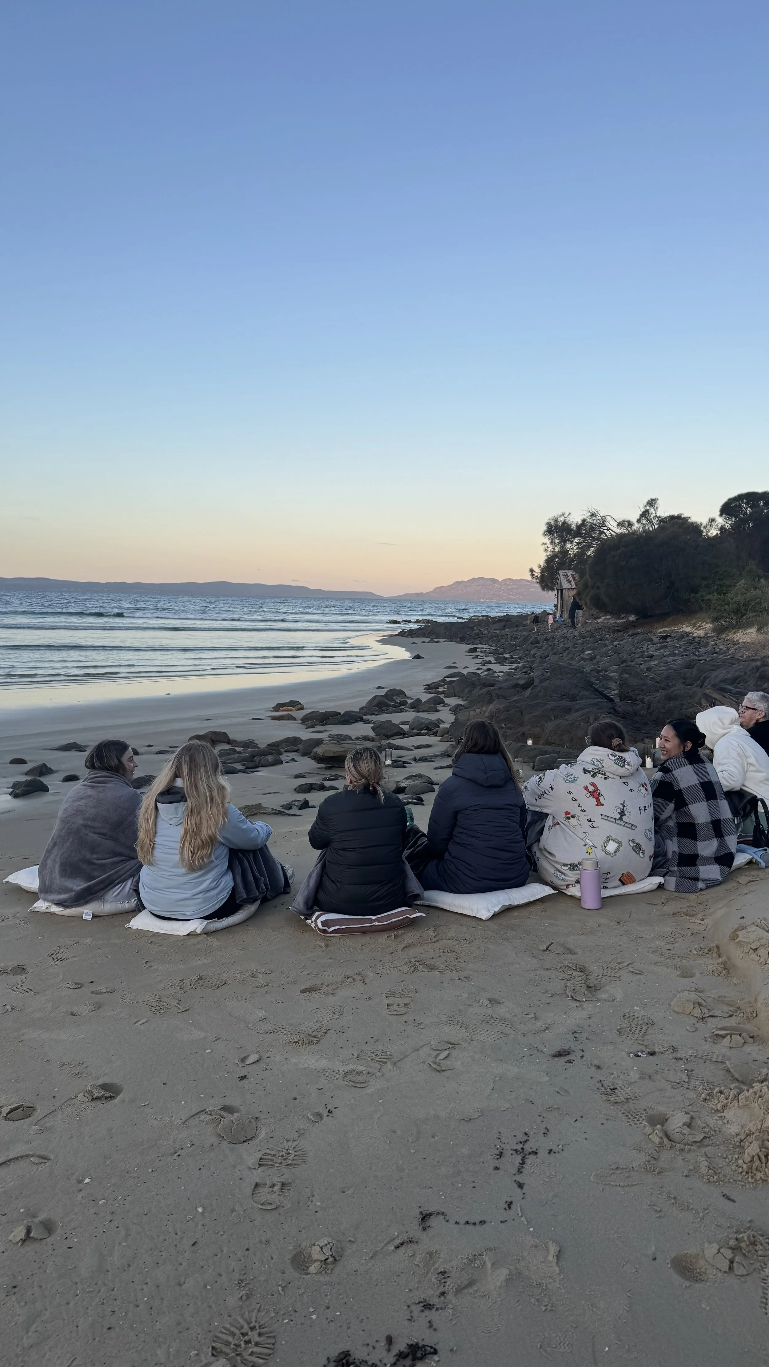 Group of people seated on cushions along a rocky beach at sunset, with waves, distant mountains, and a small beach hut.