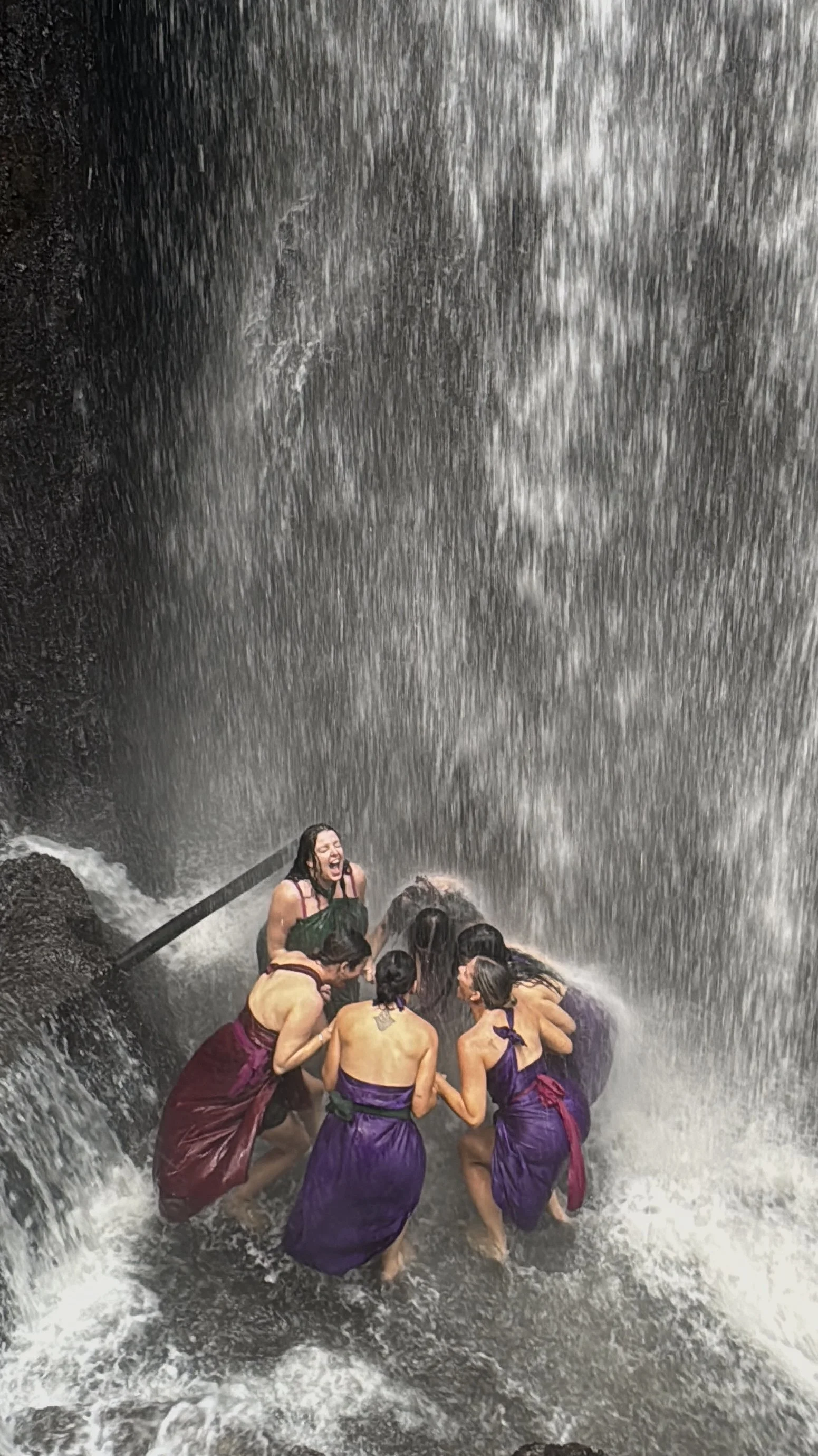 Group of people in purple sarongs standing beneath a powerful waterfall