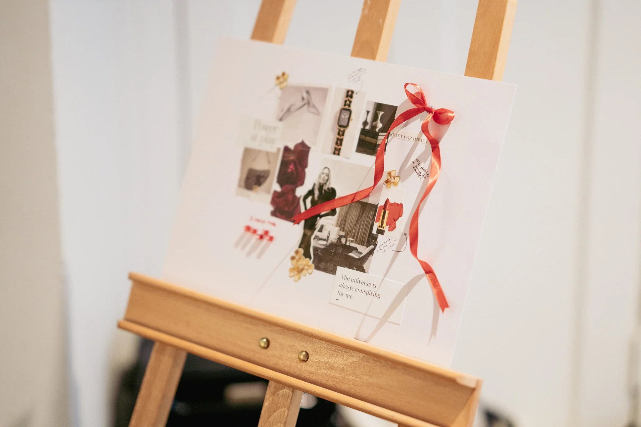 Fashion mood board with magazine clippings, jewelry, lipstick, flowers, and a red ribbon displayed on a wooden easel