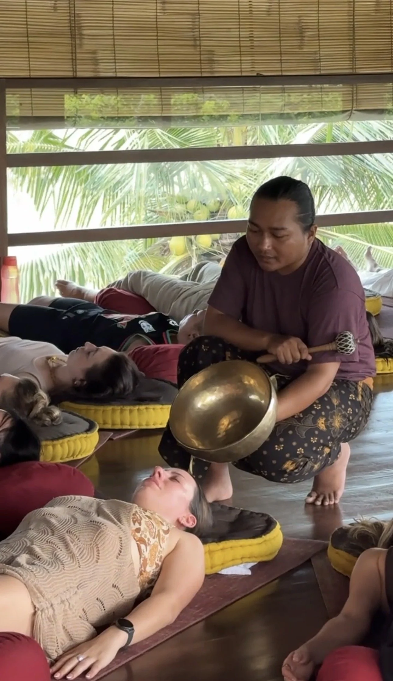 Group sound healing session with participants lying on cushions as a practitioner plays a singing bowl in a tropical room