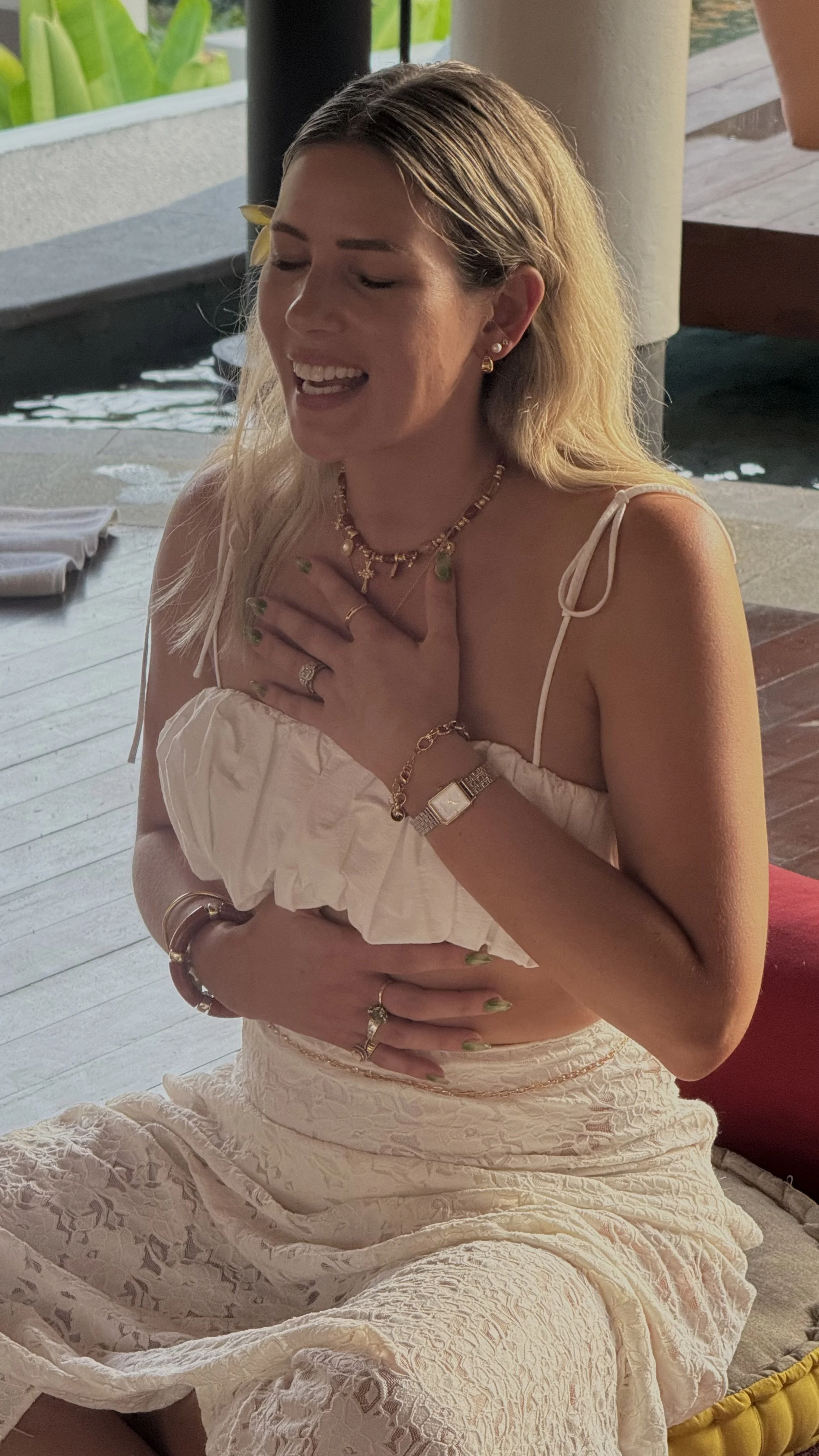 Smiling woman with blonde hair wearing a white dress and jewelry outdoors.