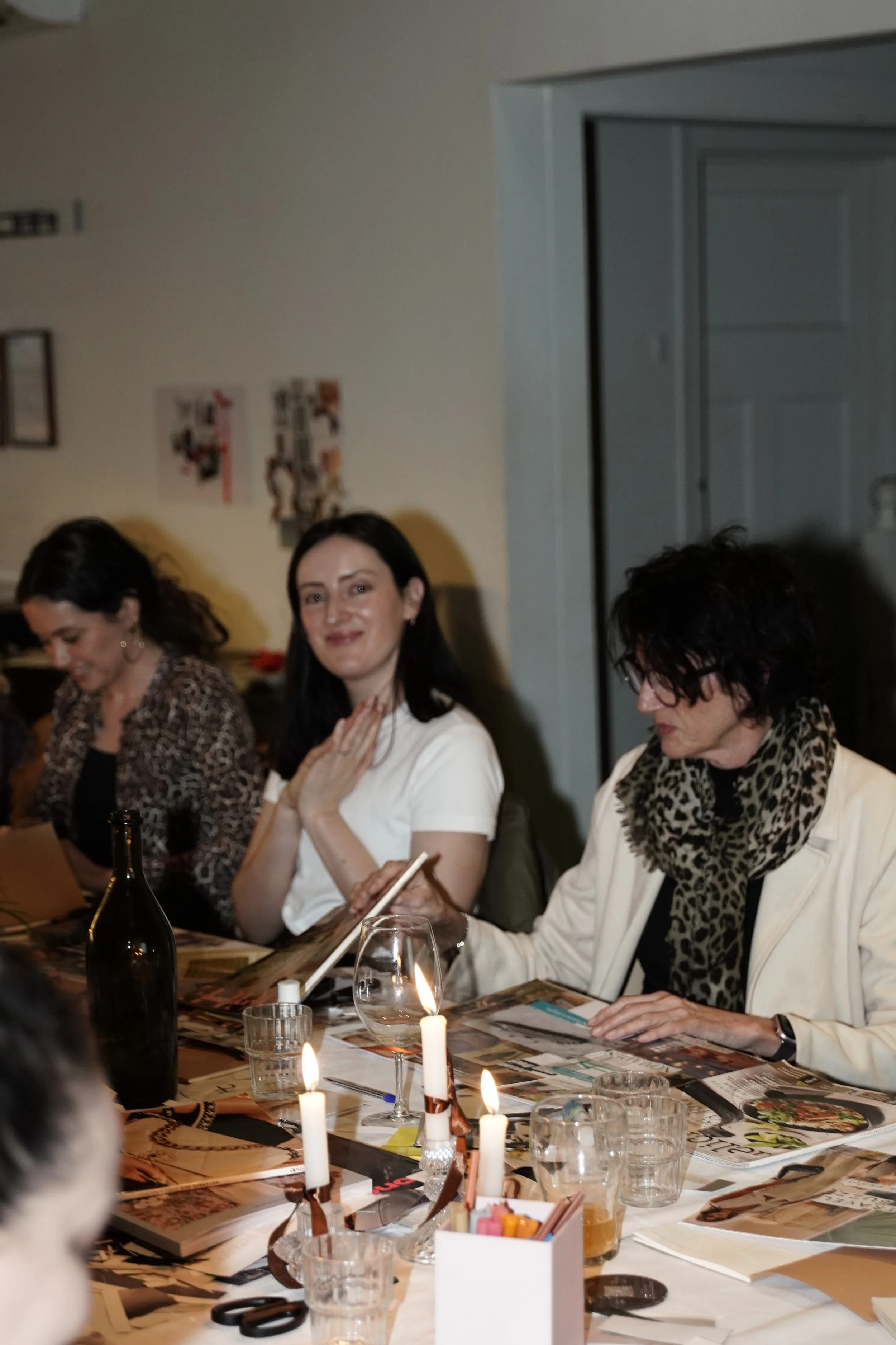 Group of women dining together at a candlelit restaurant table with wine, glasses, menus, and art supplies