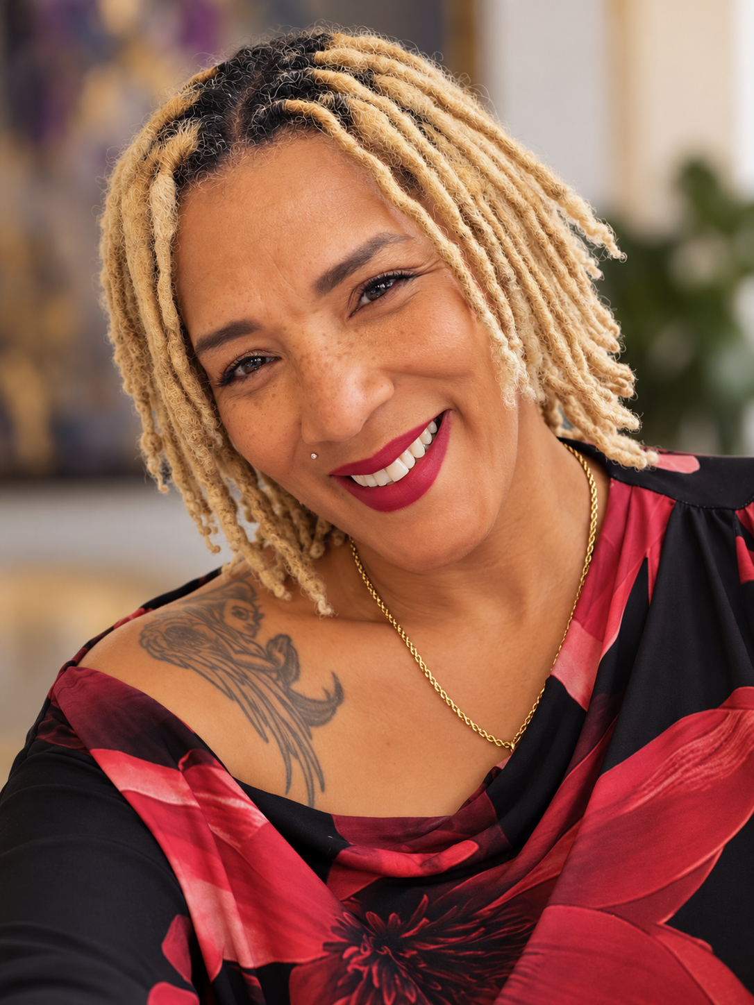 Close-up portrait of a woman with blonde dreadlocks, red lipstick, and a nose piercing, smiling at the camera. She wears a black and red floral top, a gold chain necklace, and has a tattoo of a lion on her shoulder.