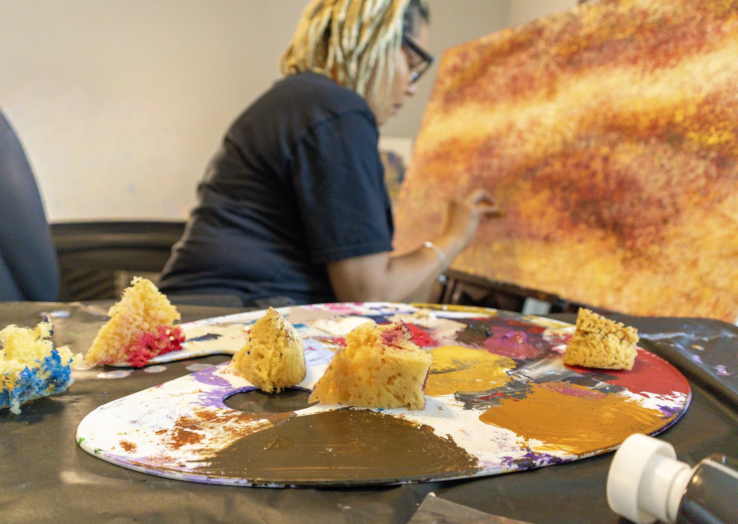 Close-up of a colorful artist's palette with paint and pieces of cake on the table, with a woman painting in the background.