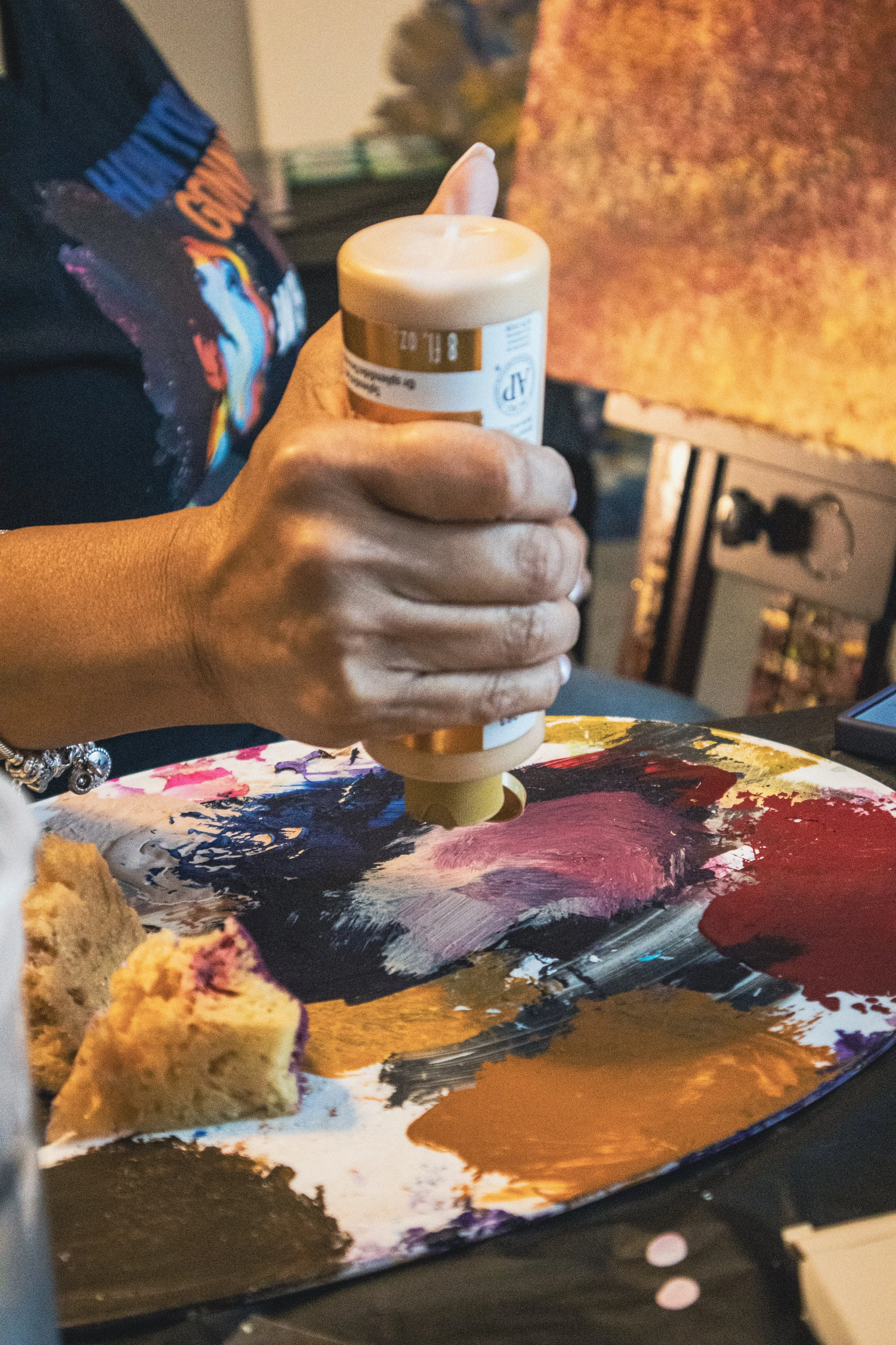 A person holding a white candle over a colorful abstract painting on a wooden surface. The painting features bold brush strokes in purple, pink, black, white, and gold. Part of a donut with pink icing and sprinkles is visible in the foreground.