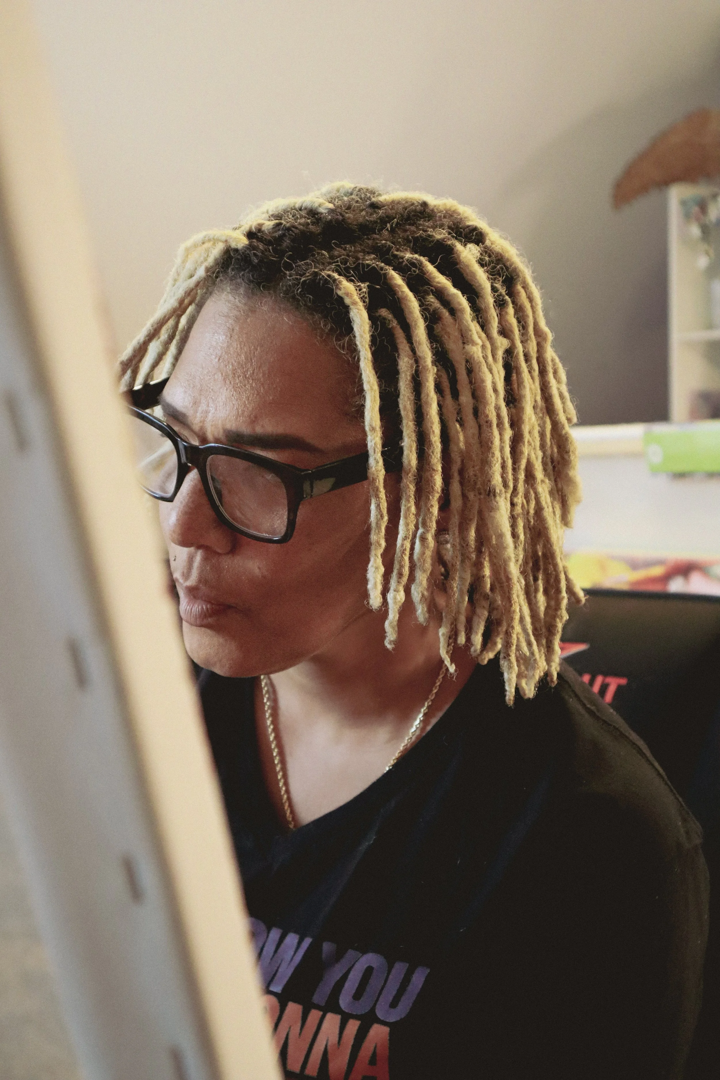 A woman with light-colored dreadlocks wearing glasses and a black T-shirt, sitting in front of a computer or tablet, focused on the screen.