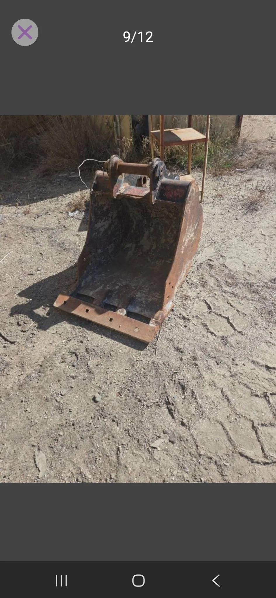 Rusty excavator bucket sitting on dirt ground outdoors with a wooden chair in background.