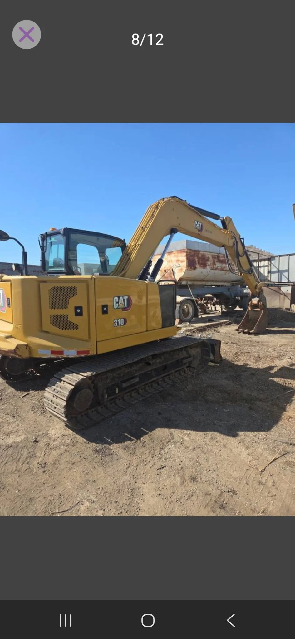 A yellow Caterpillar excavator with black tracks at a construction site, lifting a rusty metal container under a clear blue sky.