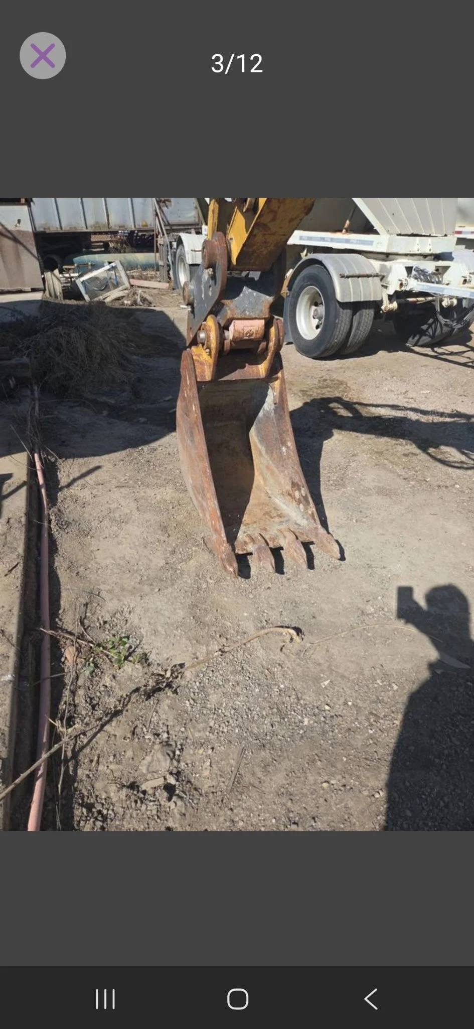 A rusty excavator bucket attachment on a construction site with dirt ground and a large truck in the background.