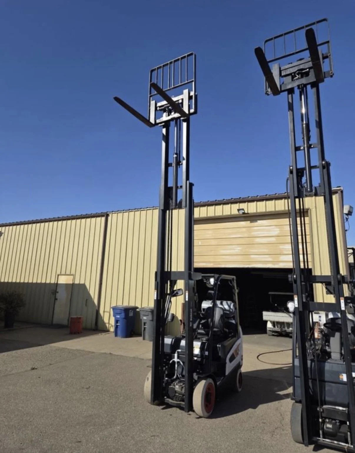 Two forklifts with extended forks for lifting pallets, parked outdoors near a beige industrial building under a clear blue sky.