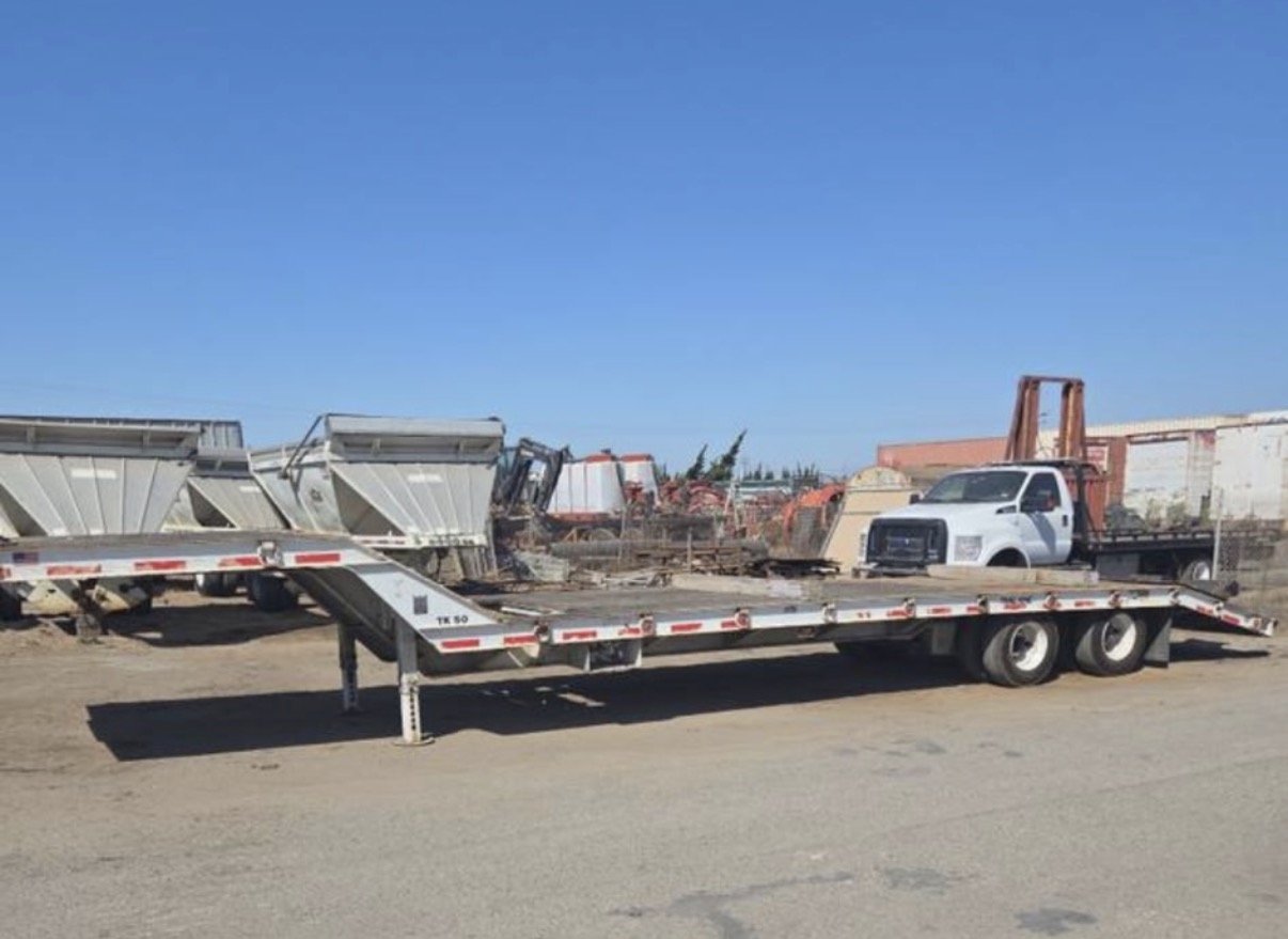 Flatbed trailer parked on dirt ground with various farm equipment and trucks in the background under a clear blue sky.