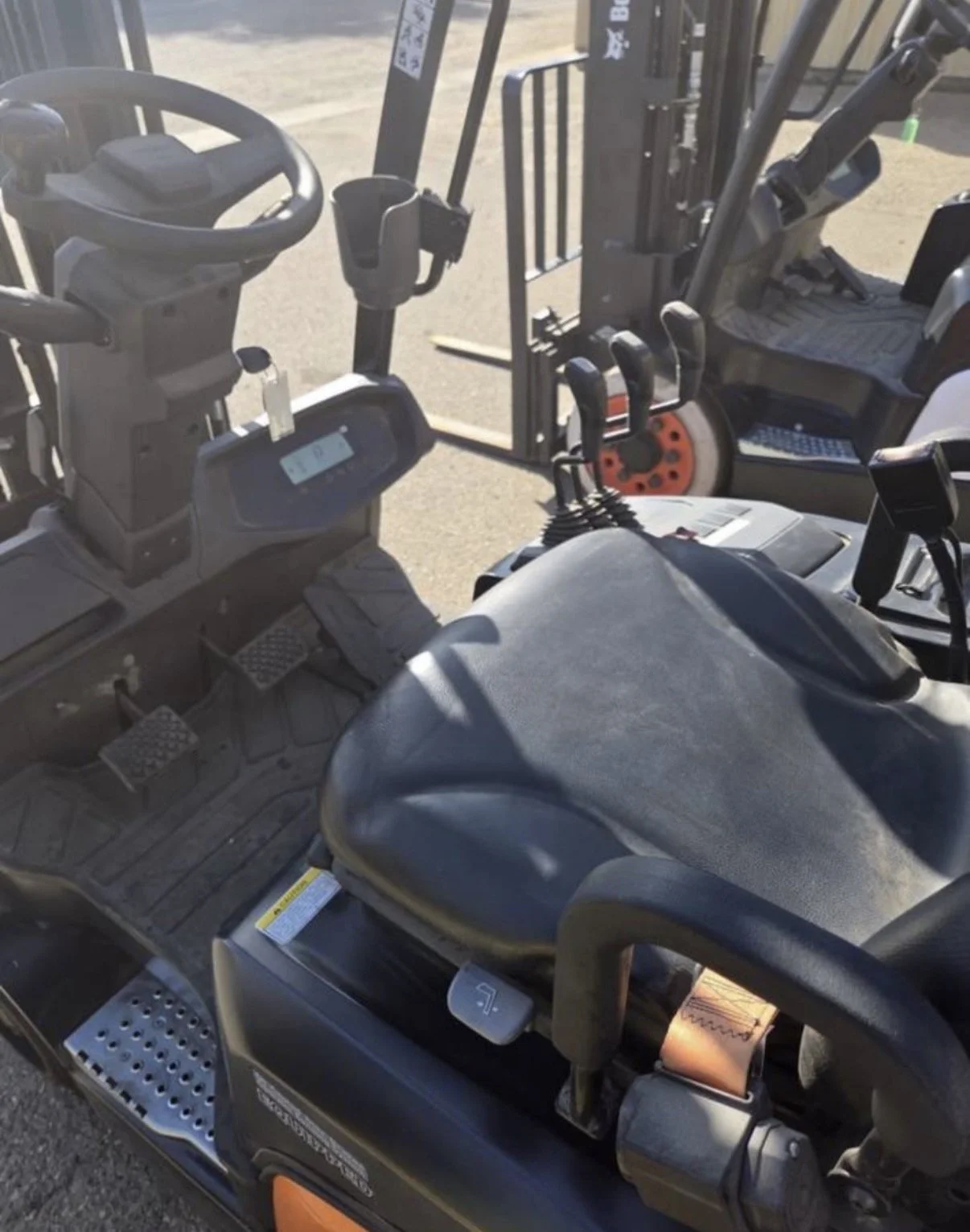 Close-up of the control panel and steering wheel of a forklift, with levers, pedals, and a seat, positioned outdoors on a concrete surface.
