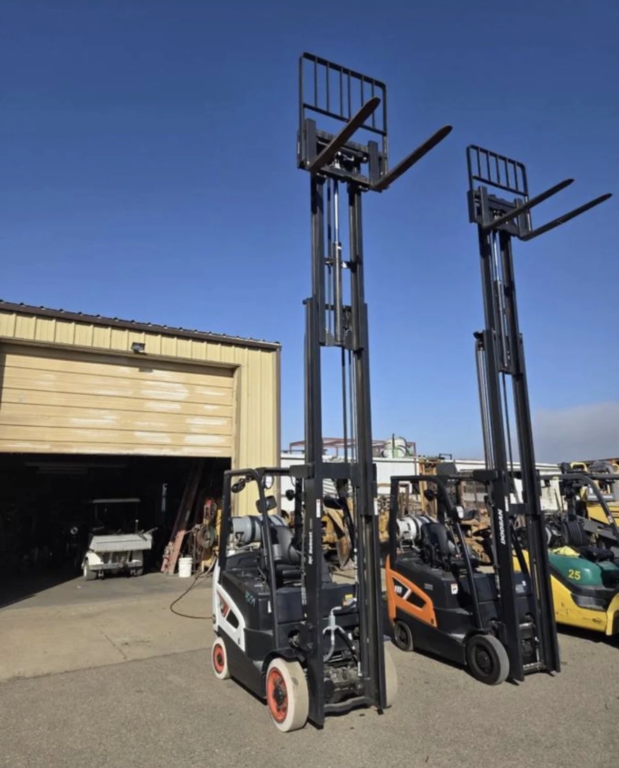 Two tall forklifts standing outdoors near a yellow building with an open garage door, with other machinery and equipment in the background under a clear blue sky.