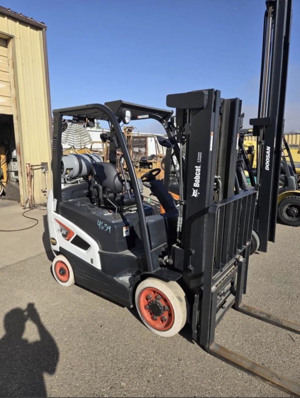 A black and white electric forklift with orange wheels parked outdoors near a yellow building, with other industrial equipment visible in the background.