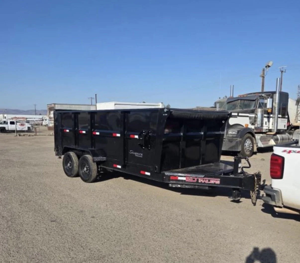 Black utility trailer attached to a white truck in a dirt lot, with other trucks and buildings in the background under a clear blue sky.
