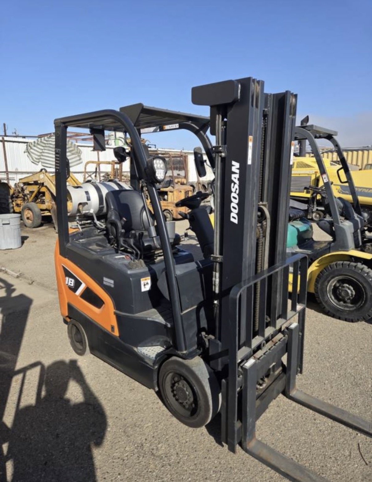 A Doosan forklift with an orange and black body and black forks, parked outdoors on a dirt surface, with industrial equipment and other forklifts in the background.
