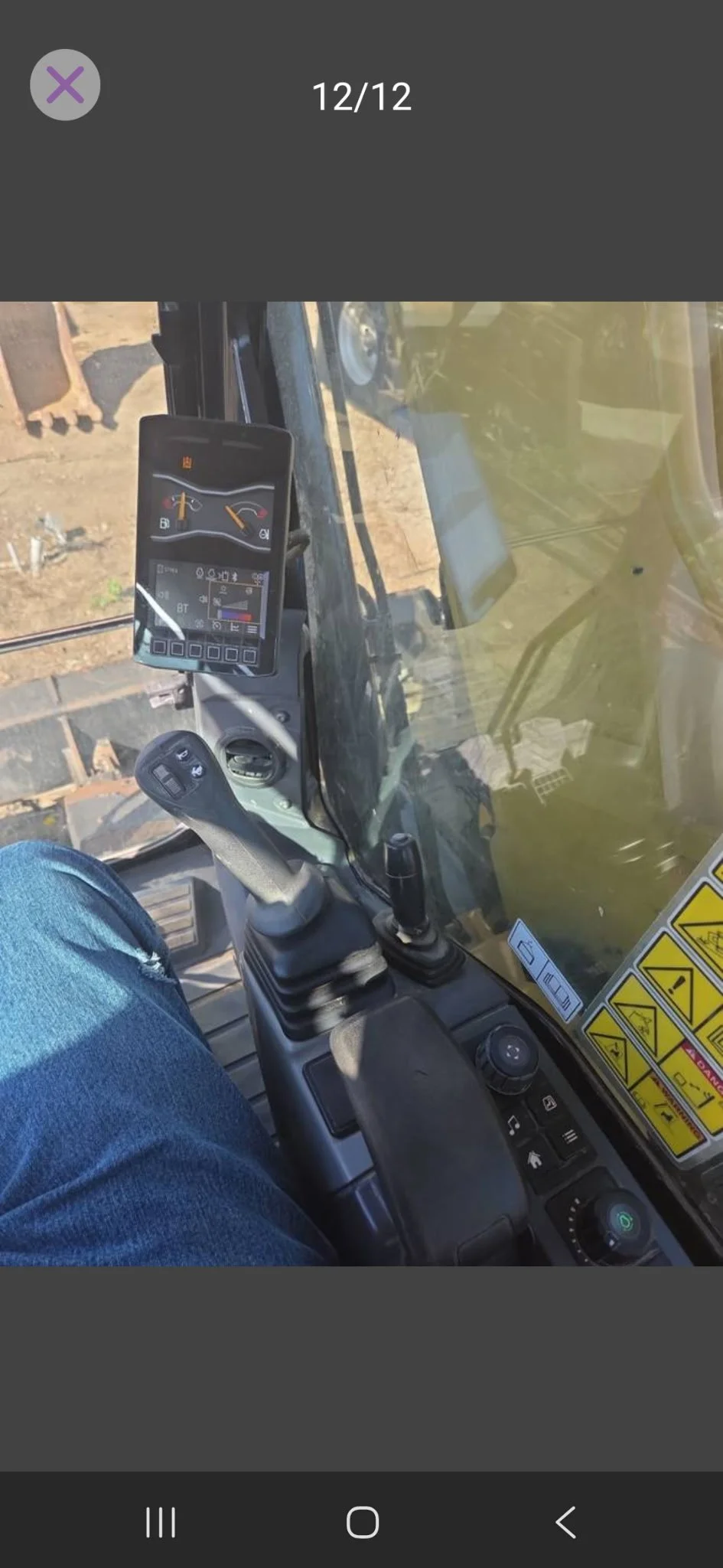 View of the control panel and interior of construction machinery, including digital displays, joysticks, and warning labels, with dirt ground visible outside.
