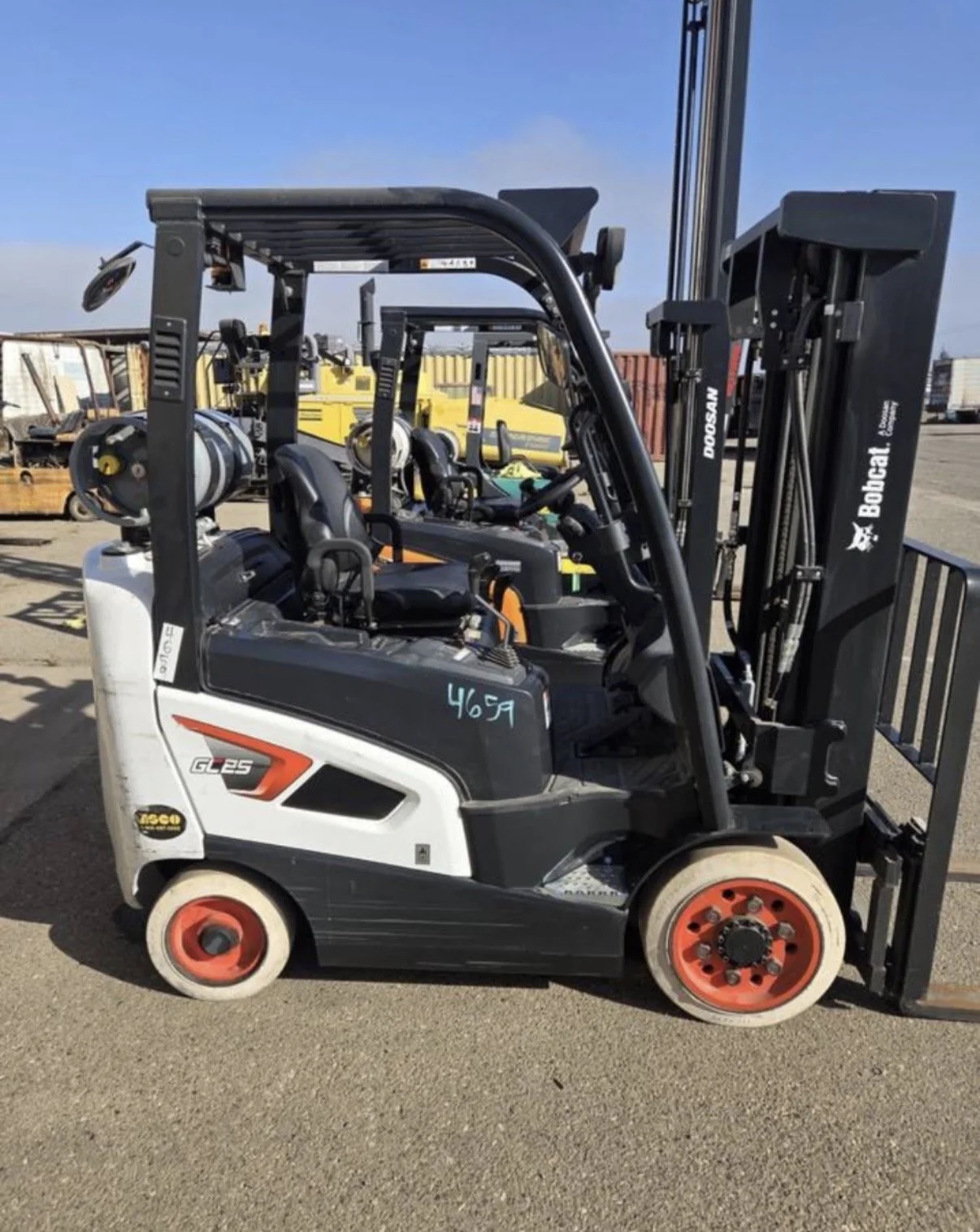 A Bobcat GL25 compact forklift with red wheels, enclosed operator seat and overhead guard, parked outdoors on a gravel surface, with storage containers and equipment in the background.