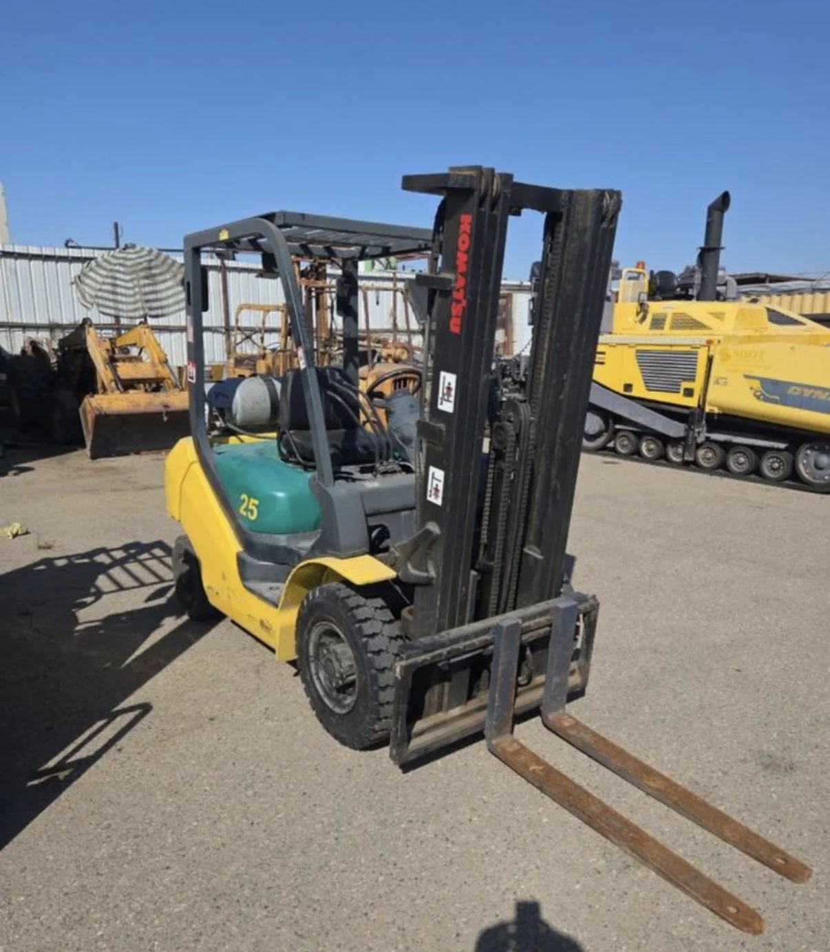 Yellow and green forklift truck parked on gravel with old construction equipment and machinery in the background under a clear blue sky.