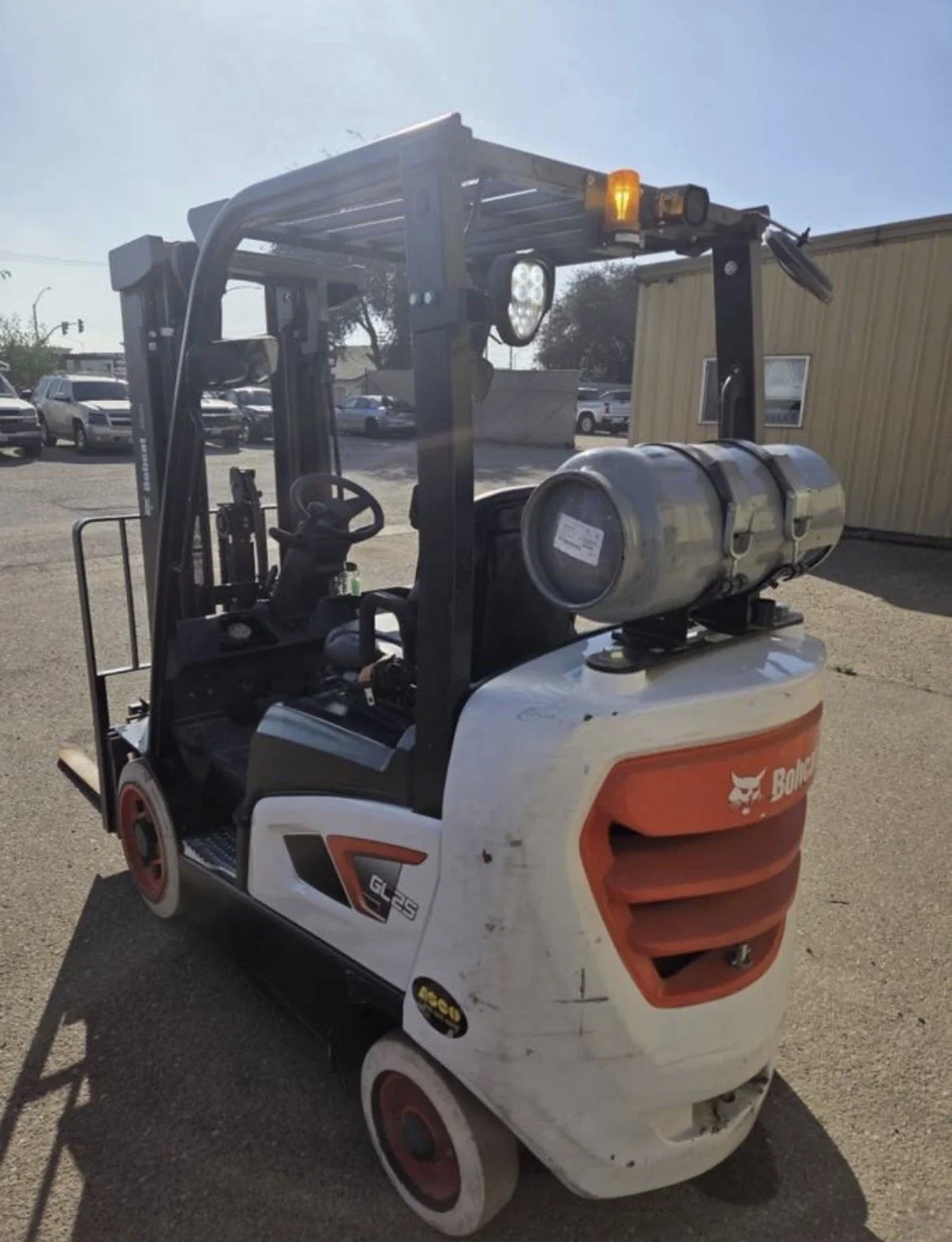 A white forklift with a propane tank on the back, parked outdoors on a sunny day, with several cars and a beige building in the background.