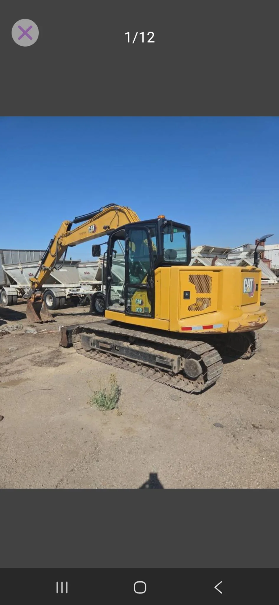 A yellow Caterpillar mini excavator with tracked wheels parked on dirt ground, with trailers in the background and a clear blue sky.